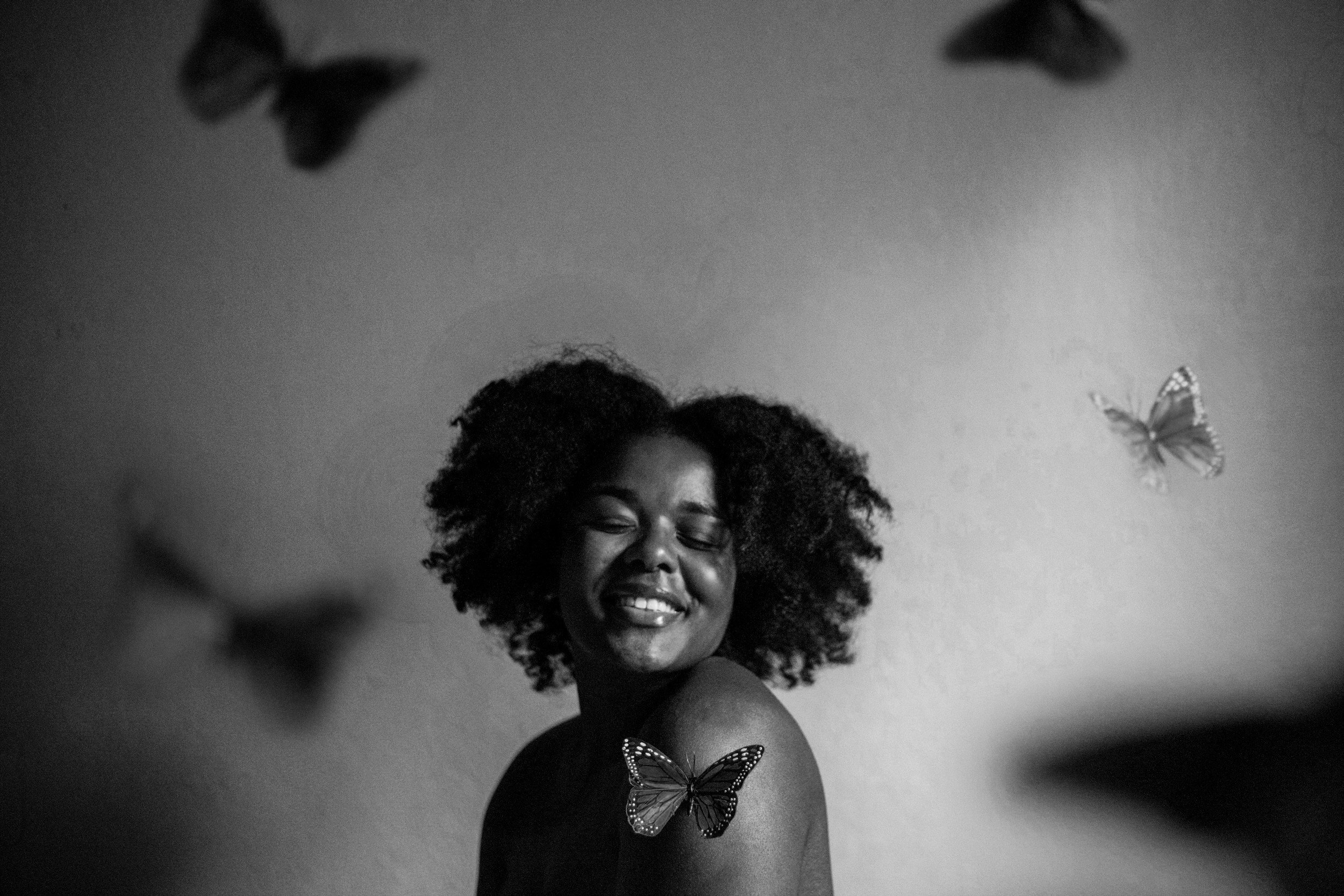 A black-and-white photo of a woman with curly hair smiling with eyes closed, surrounded by butterflies on the wall and on her shoulder.