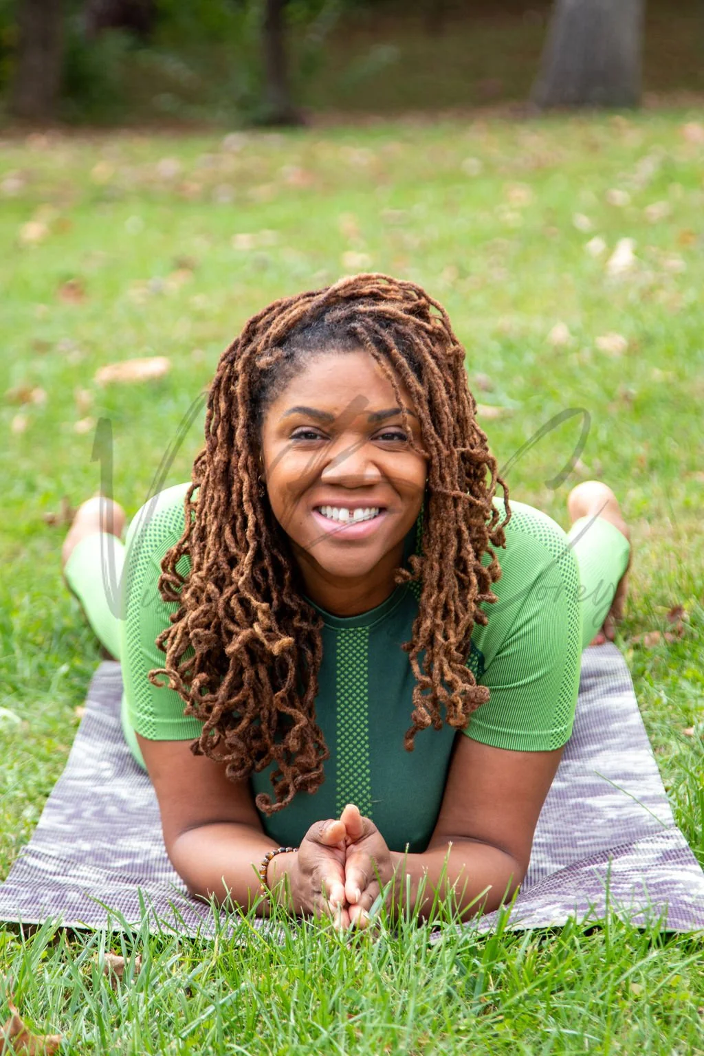 A woman with long dreadlocks practicing yoga outdoors on a grassy field, lying on her stomach with her hands clasped and smiling at the camera.
