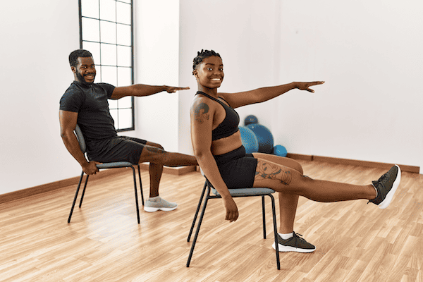 Two people in a fitness studio seated on chairs, extending their arms and legs in a stretching exercise.