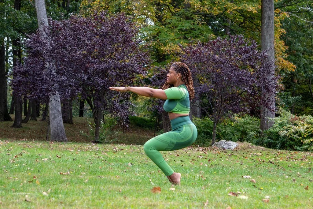 Woman practicing yoga outdoors in a park with trees and fallen leaves, wearing a green yoga outfit, in a squat pose with arms extended forward.