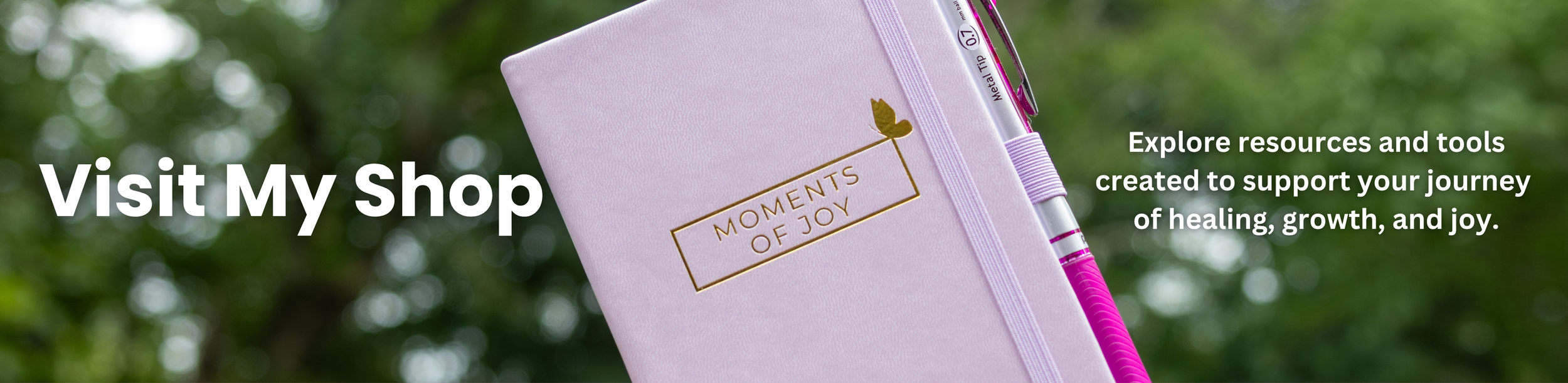 A light pink fabric notebook with gold embossed text that reads "Moments of Joy," placed outdoors against a background of blurred green foliage. The notebook has a light pink elastic band and a pink pen clipped to it.