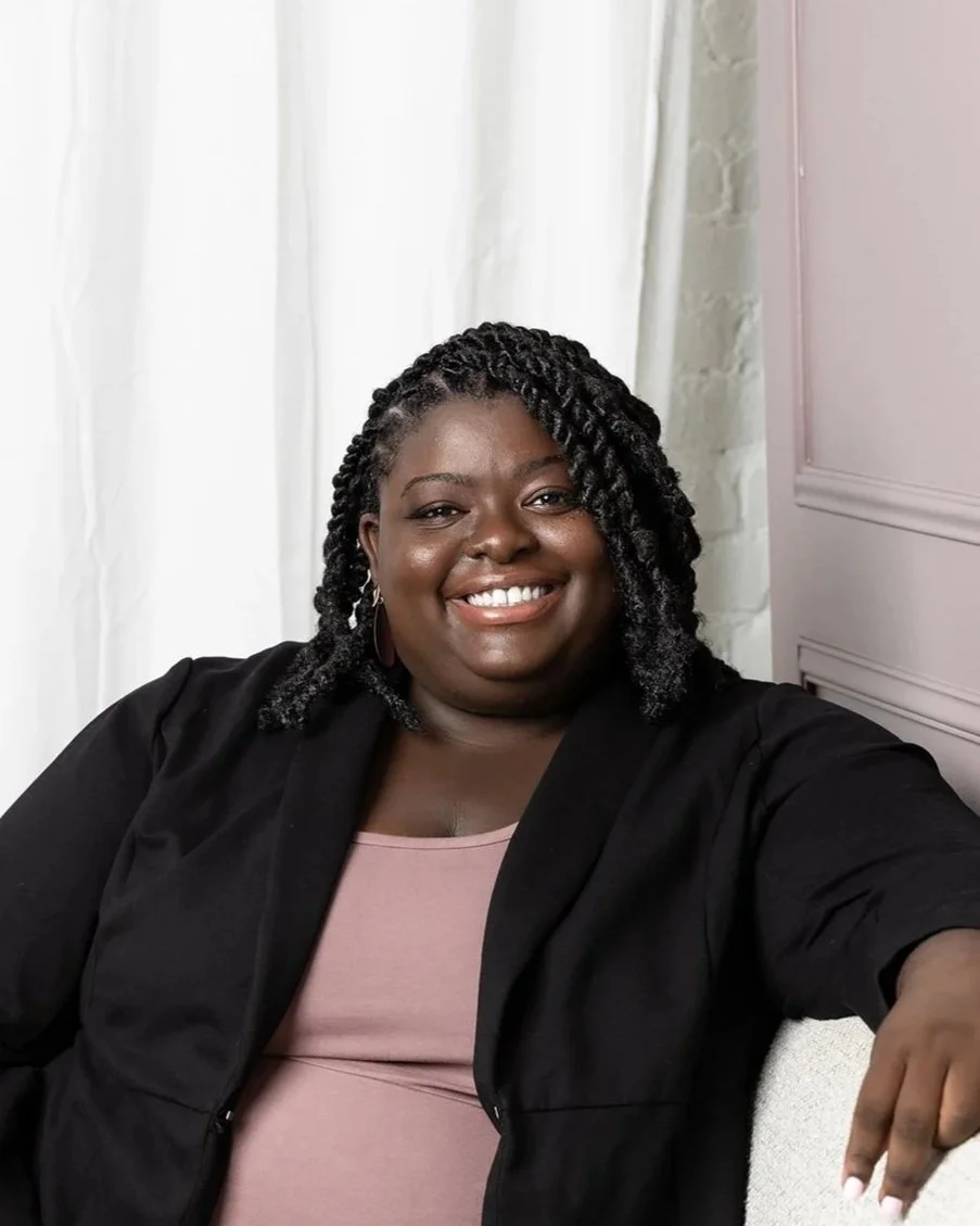 A woman with dark skin and black twisted hair smiles while sitting on a light-colored sofa in a room with white curtains and pinkish wall paneling.