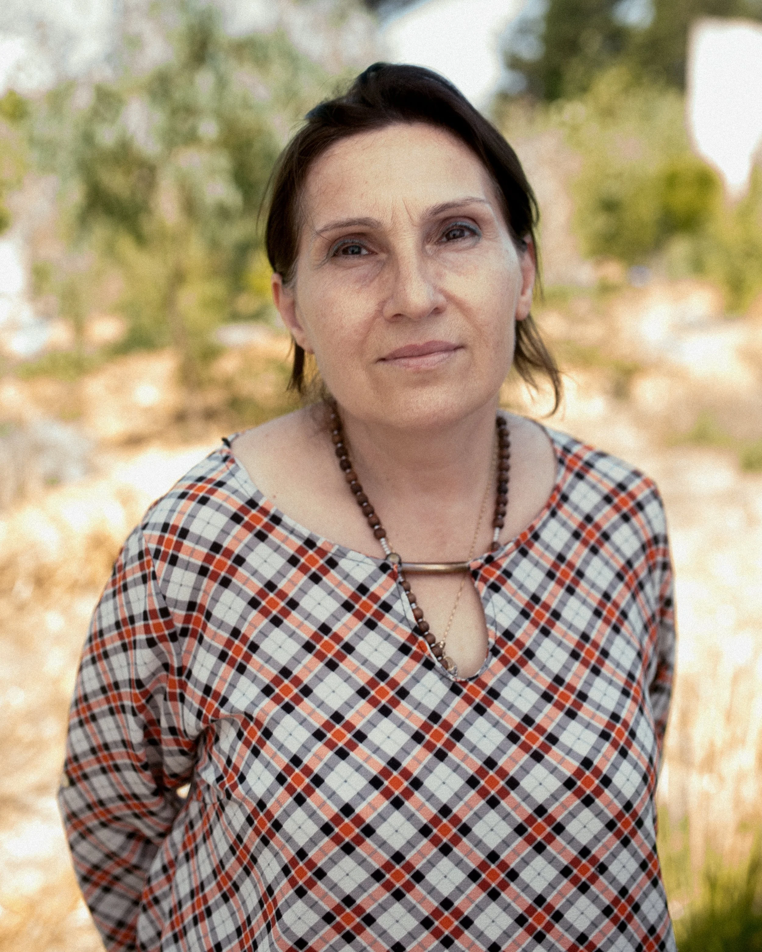 A woman wearing a plaid dress and necklace, standing outdoors with a blurred natural background.