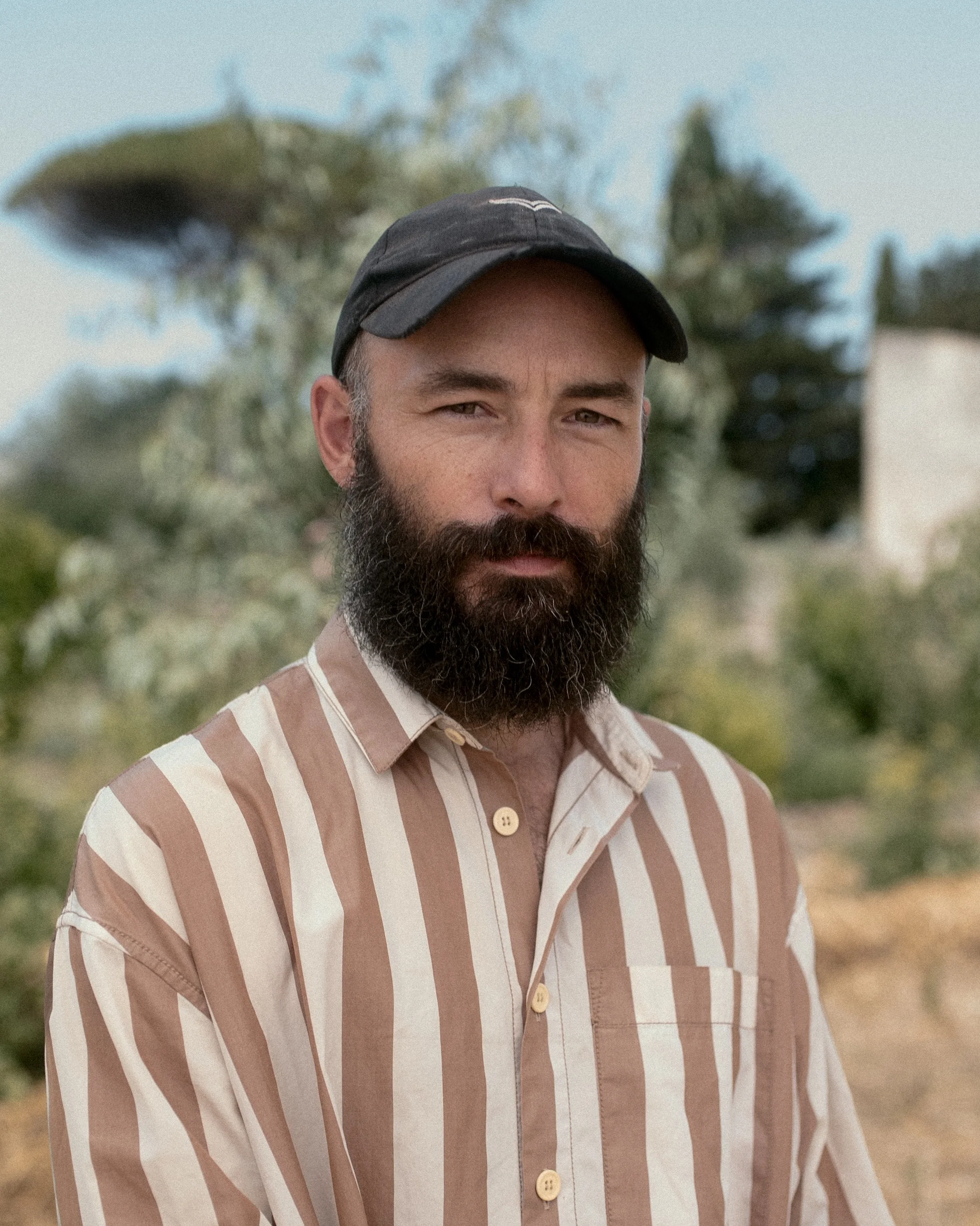 Man with a beard and cap wearing a striped shirt standing outdoors with trees in the background.
