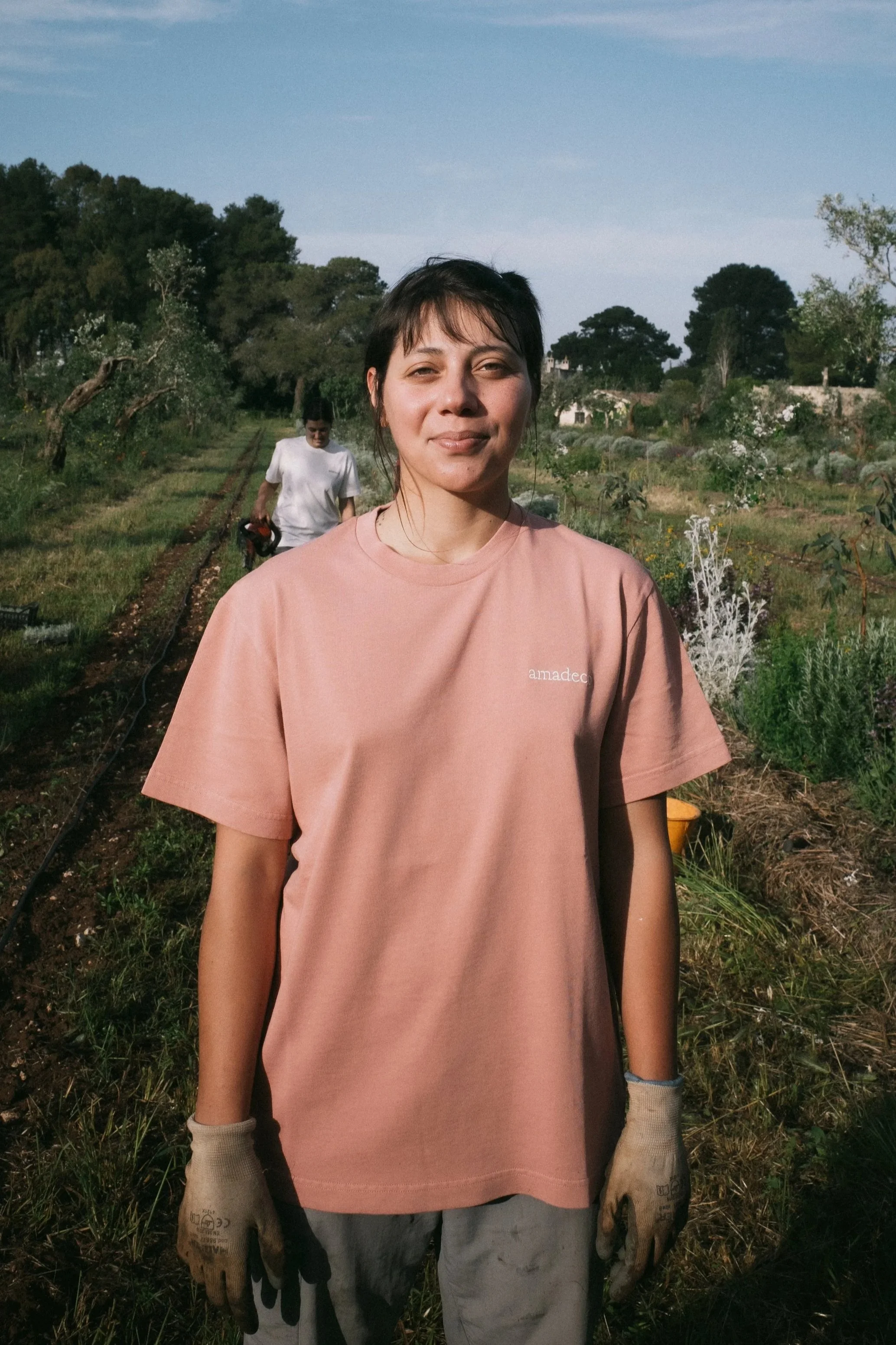 A woman standing in a garden, wearing a pink t-shirt, gardening gloves, and work clothes, with another person working in the background.