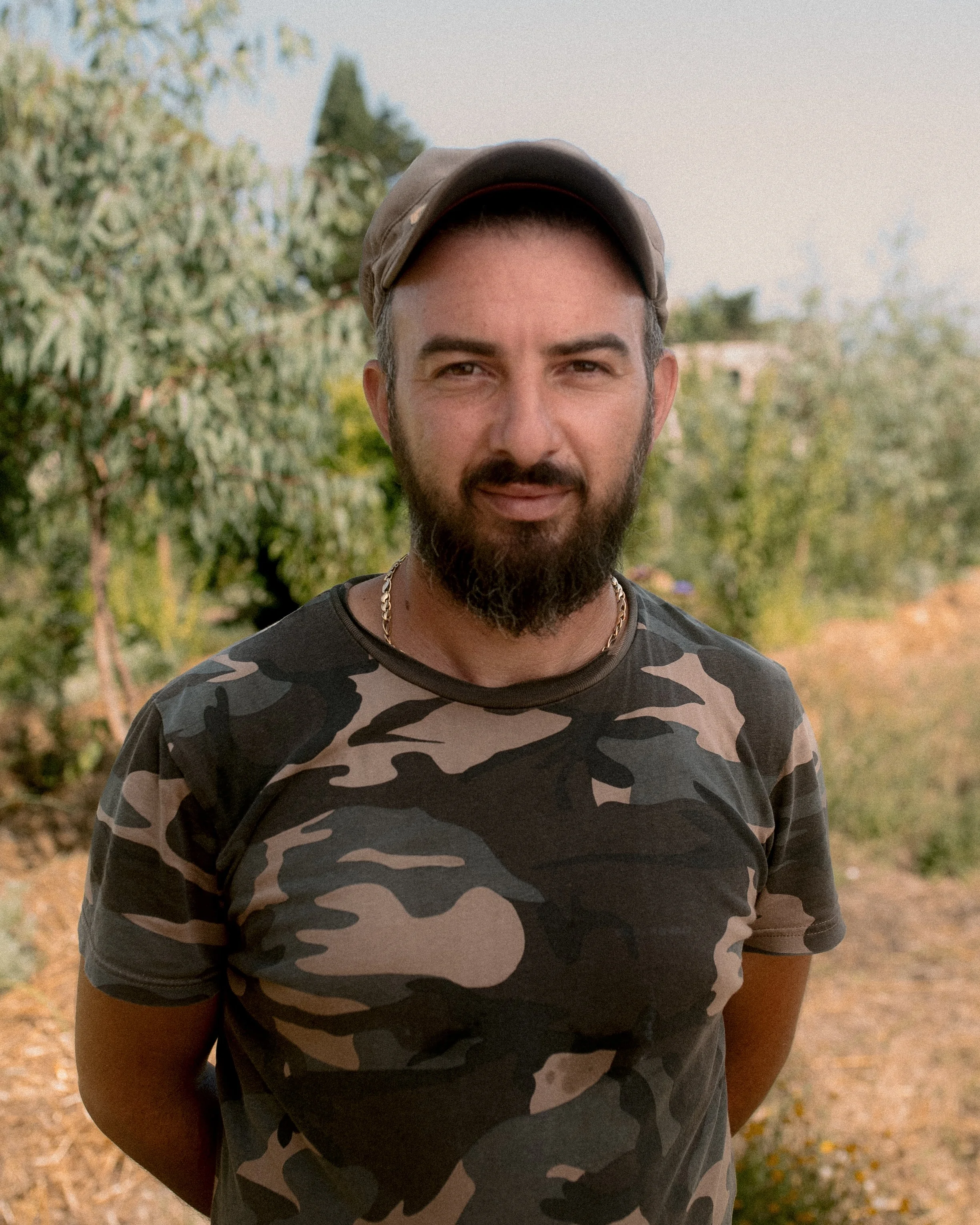 Man in camouflage shirt and cap standing outdoors with trees in the background.