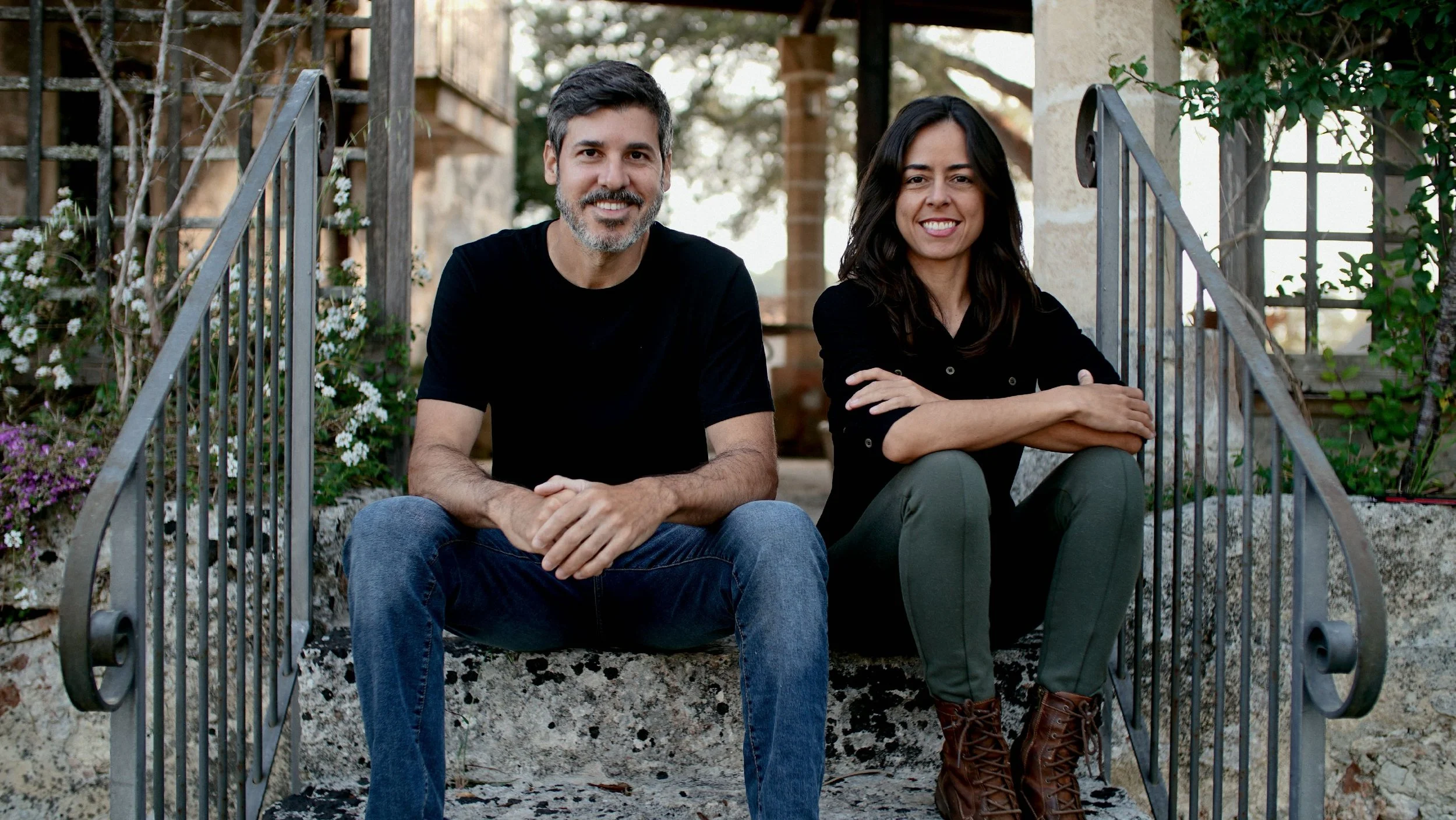 A middle-aged man with gray hair and beard, wearing a black t-shirt, and middle-aged woman with dark hair, wearing a black top, sitting on steps outside surrounded by greenery and flowers.