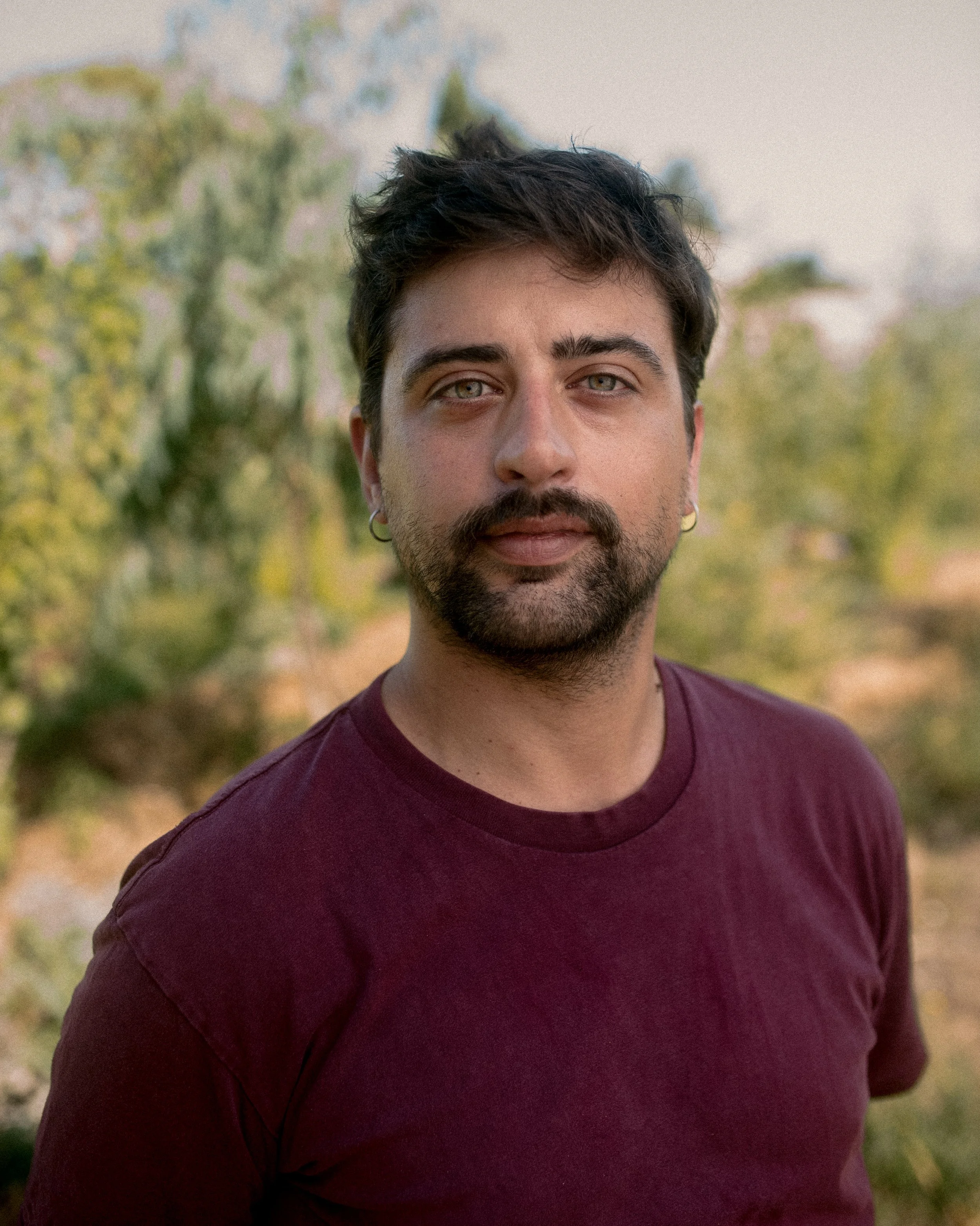 Portrait of a man with short hair and facial hair, wearing a maroon t-shirt, standing outdoors with blurred greenery in the background.