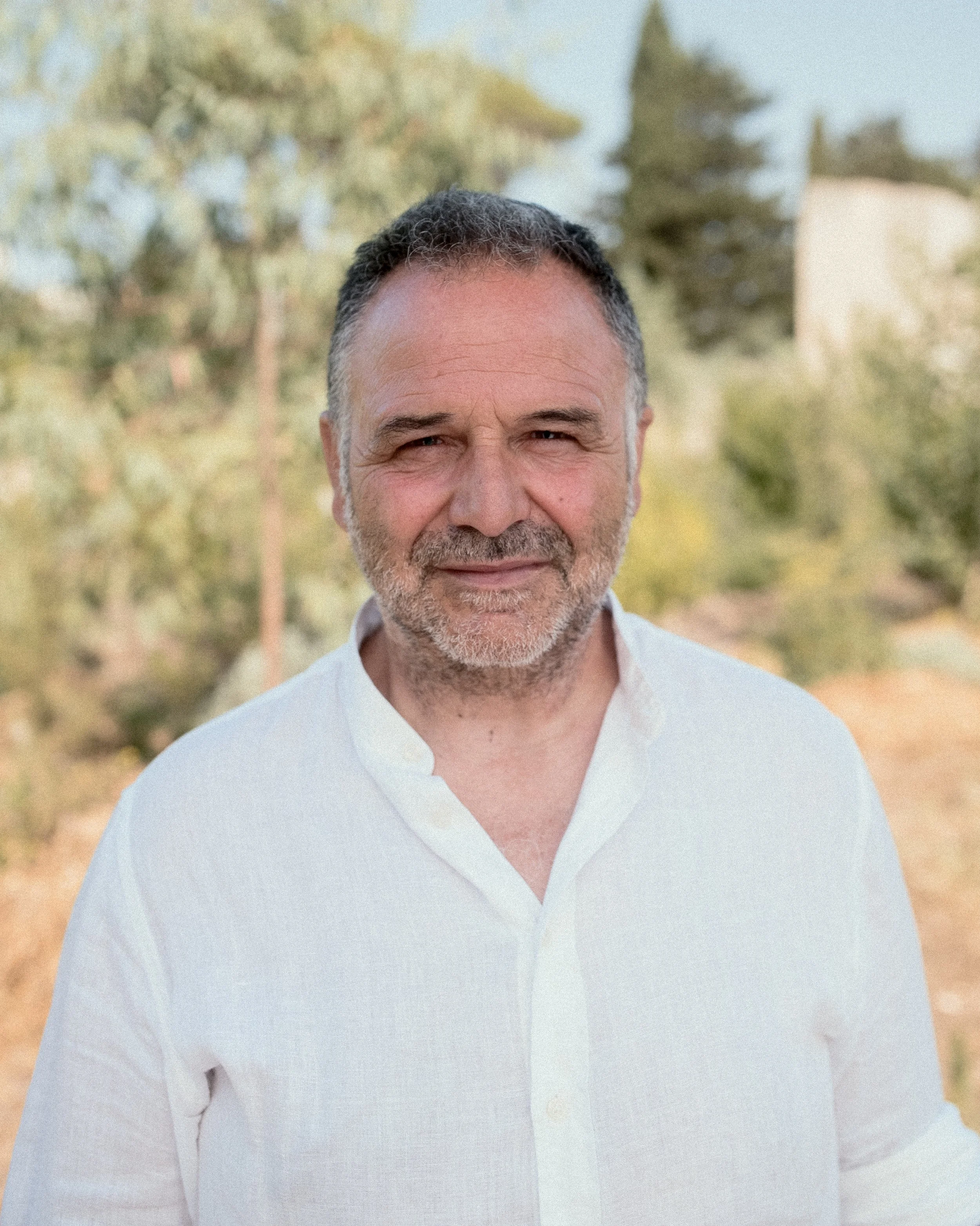 Man in a white shirt standing outdoors with blurred trees in the background.