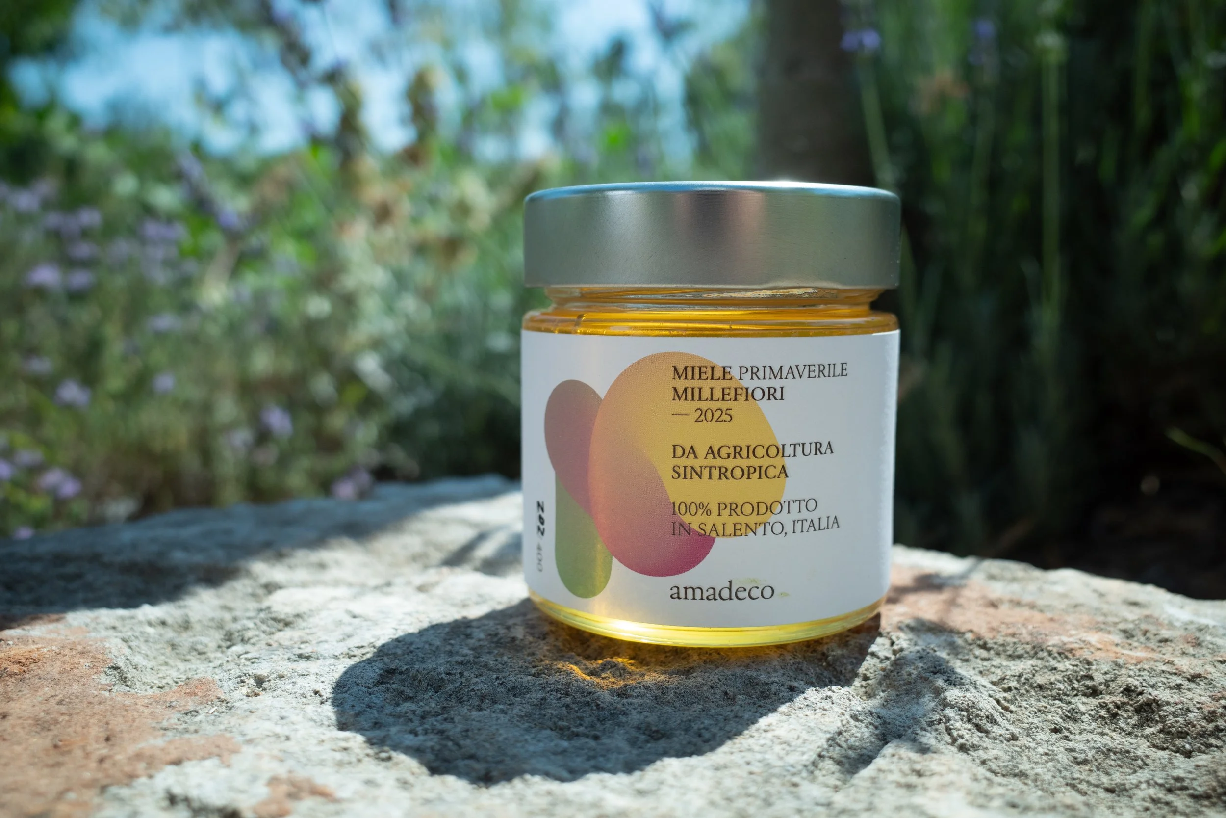 A jar of honey sitting on a stone surface outdoors, with a blurred green background of trees and plants.