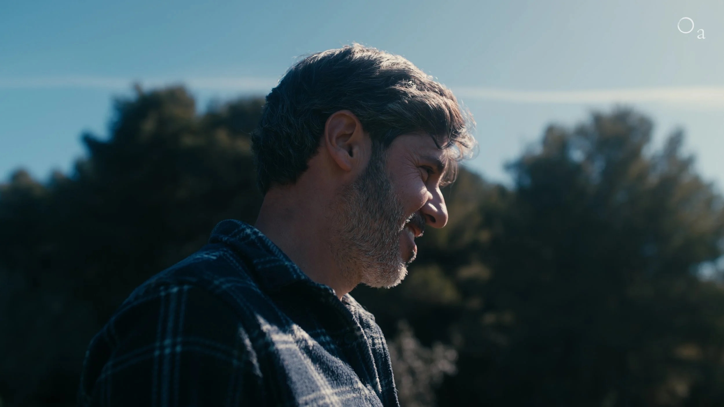 Side view of a man in a plaid shirt outdoors with trees in the background on a sunny day.