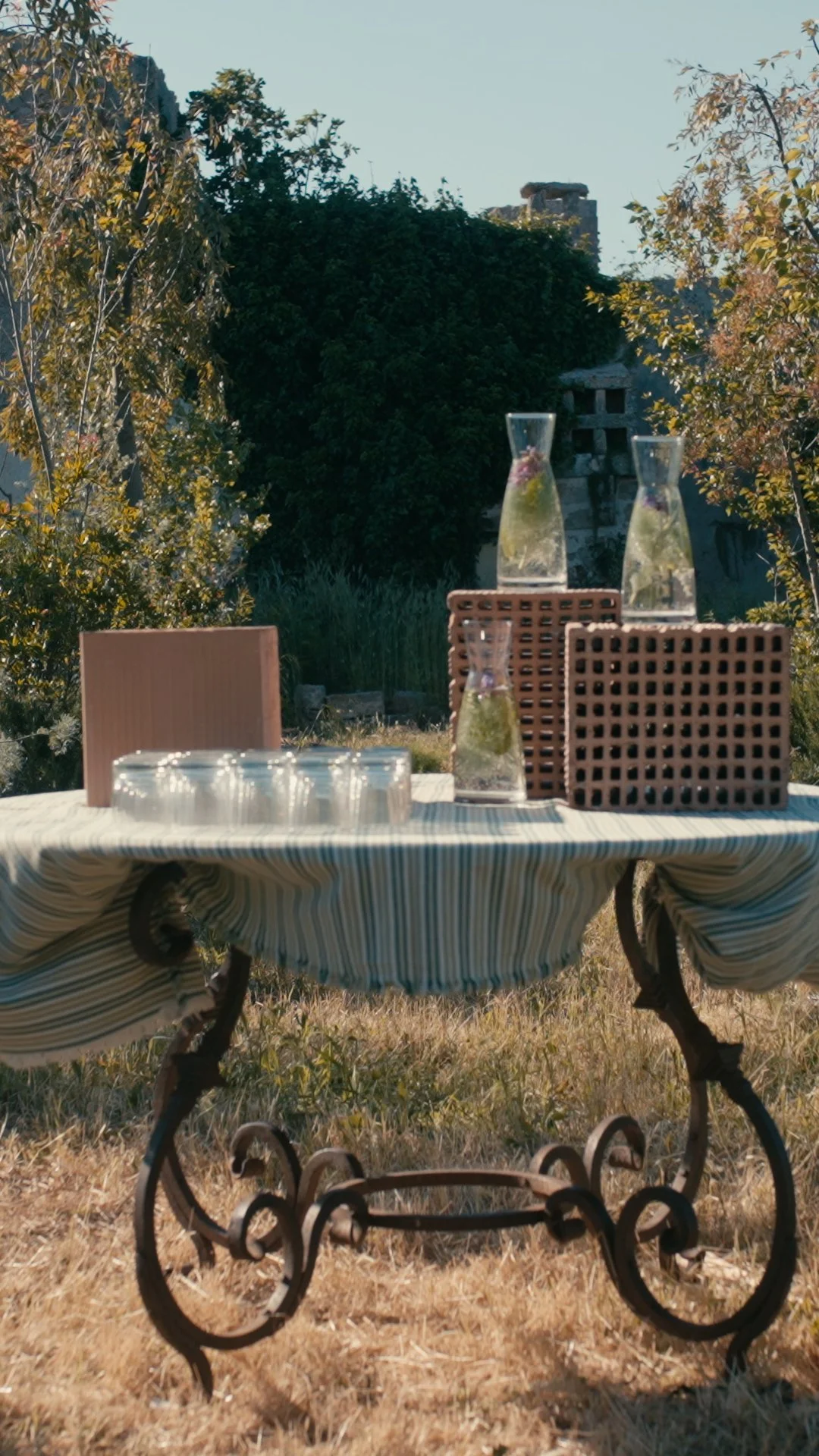 Outdoor table set with three glass carafes filled with water and floating flowers, four upside-down glasses, a pink box, and two brown lattice-patterned containers, on a striped tablecloth, in a backyard with trees and a stone wall in the background.