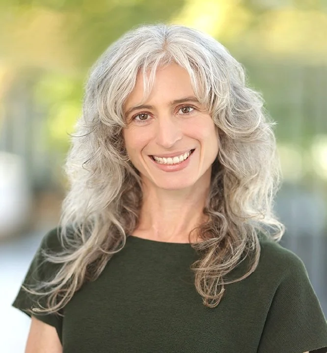 A smiling woman with long, curly gray hair stands in front of a plain white wall, wearing a sleeveless black top and a green bead necklace, with pink flowers partially visible behind her.