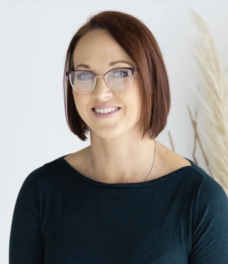 A woman with shoulder-length red hair and glasses, smiling and wearing a black top, standing in front of a white background with dried decorative plants.
