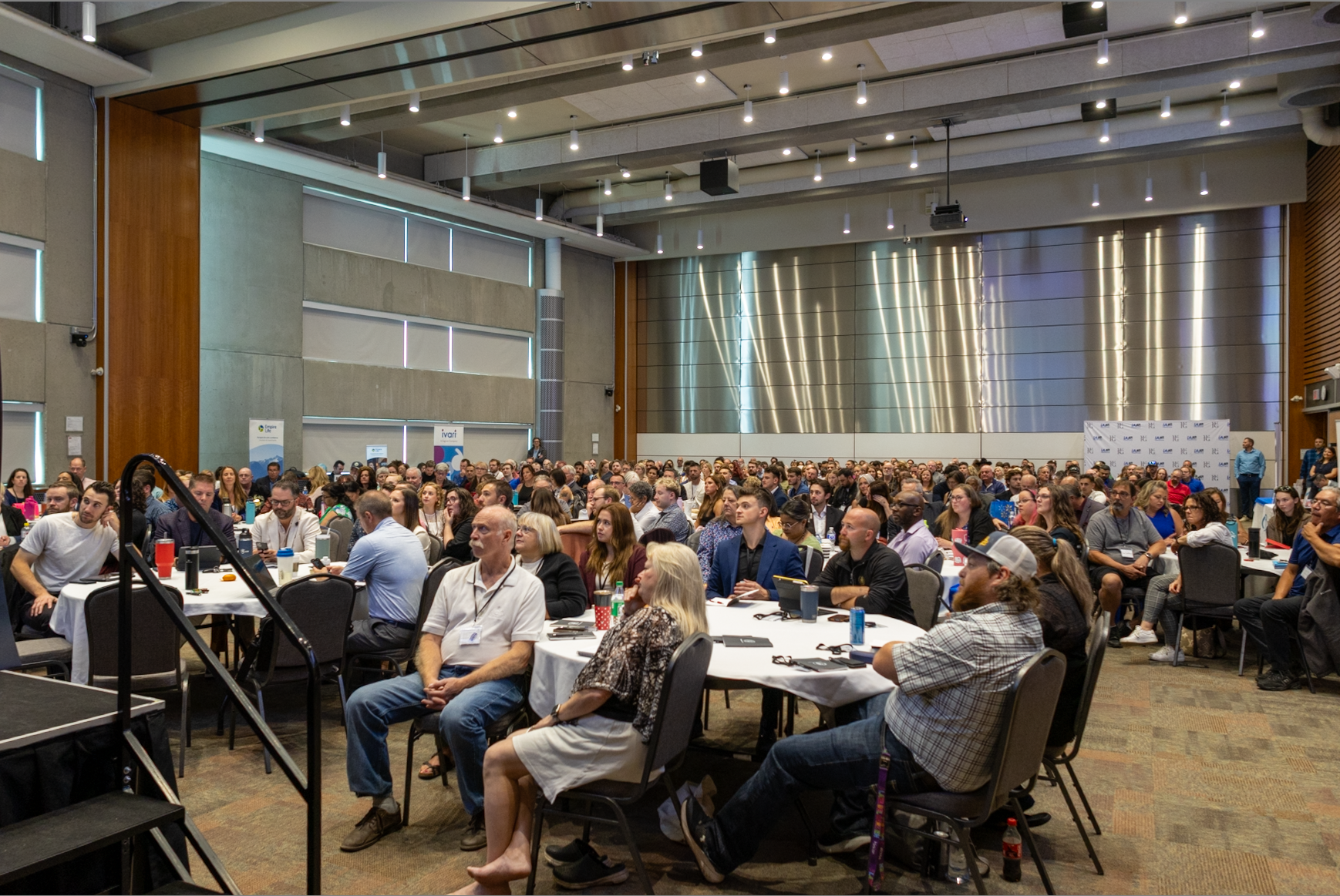 A large conference room filled with numerous attendees seated at round tables, facing a stage at the front. The room has high ceilings with modern lighting, wood paneling, and a metallic wall at the back.