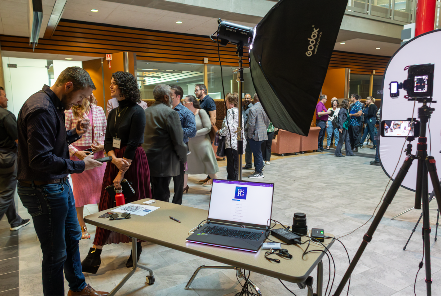 A busy indoor event space with a group of people waiting in line and engaging in conversation. A table with a laptop displaying 'WG' and equipment for photography or video recording, including a large light and camera setup, is present in the foreground.