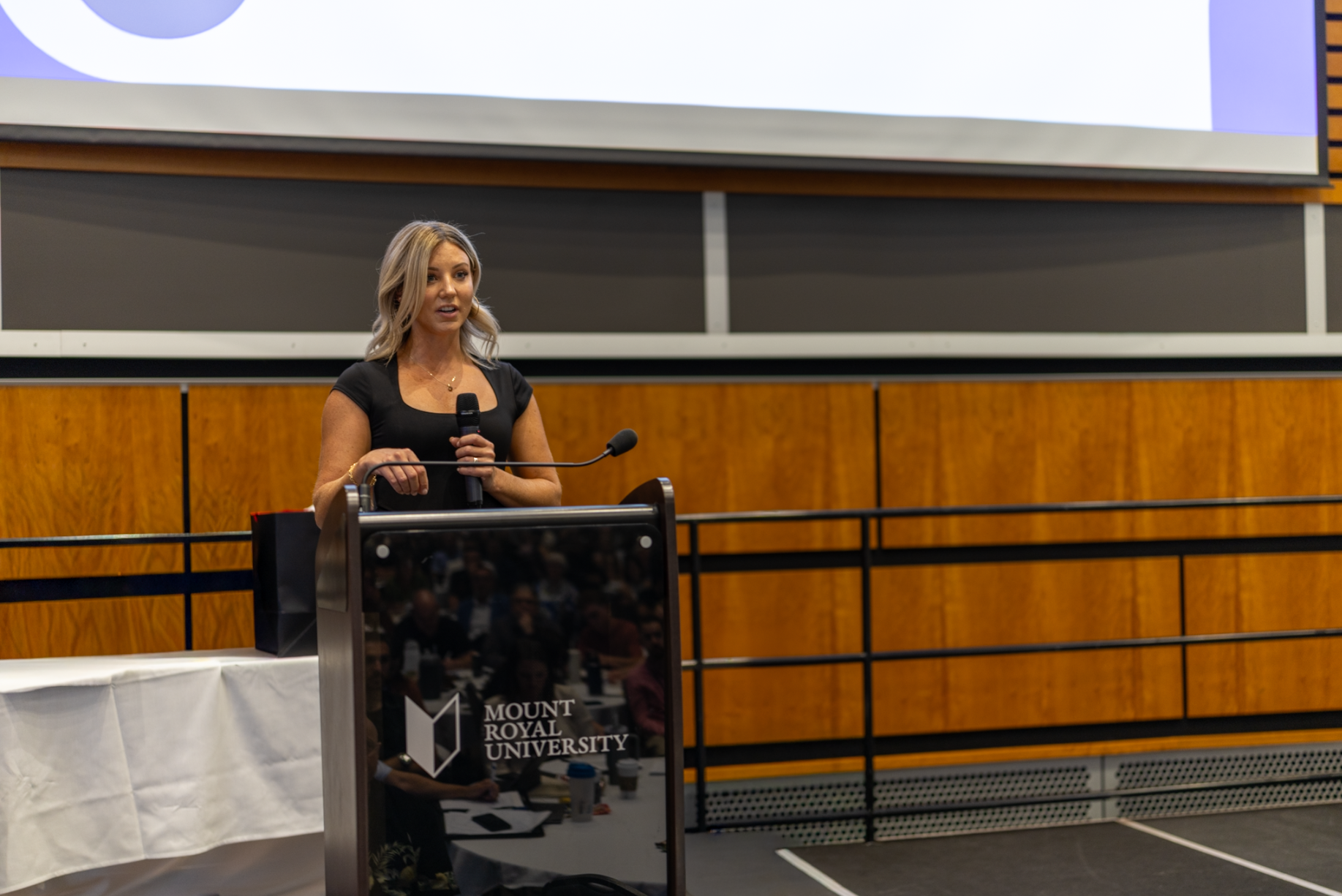 A woman giving a presentation at Mount Royal University, standing behind a podium with a microphone, in a lecture hall with wood-paneled walls and a large projection screen overhead.