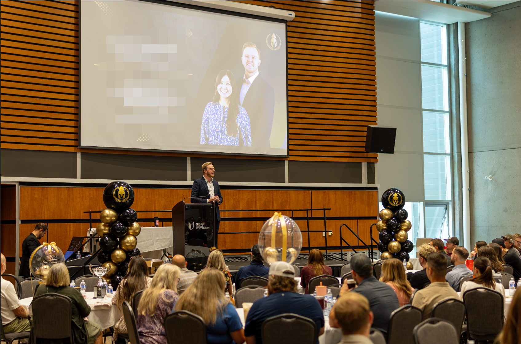 A man in a suit gives a speech at a conference, standing behind a podium decorated with balloons in black and gold. The large screen behind him shows a photo of a smiling man and woman. The event is at Mount Royal University, with seated attendees watching the speaker in a large, modern auditorium.