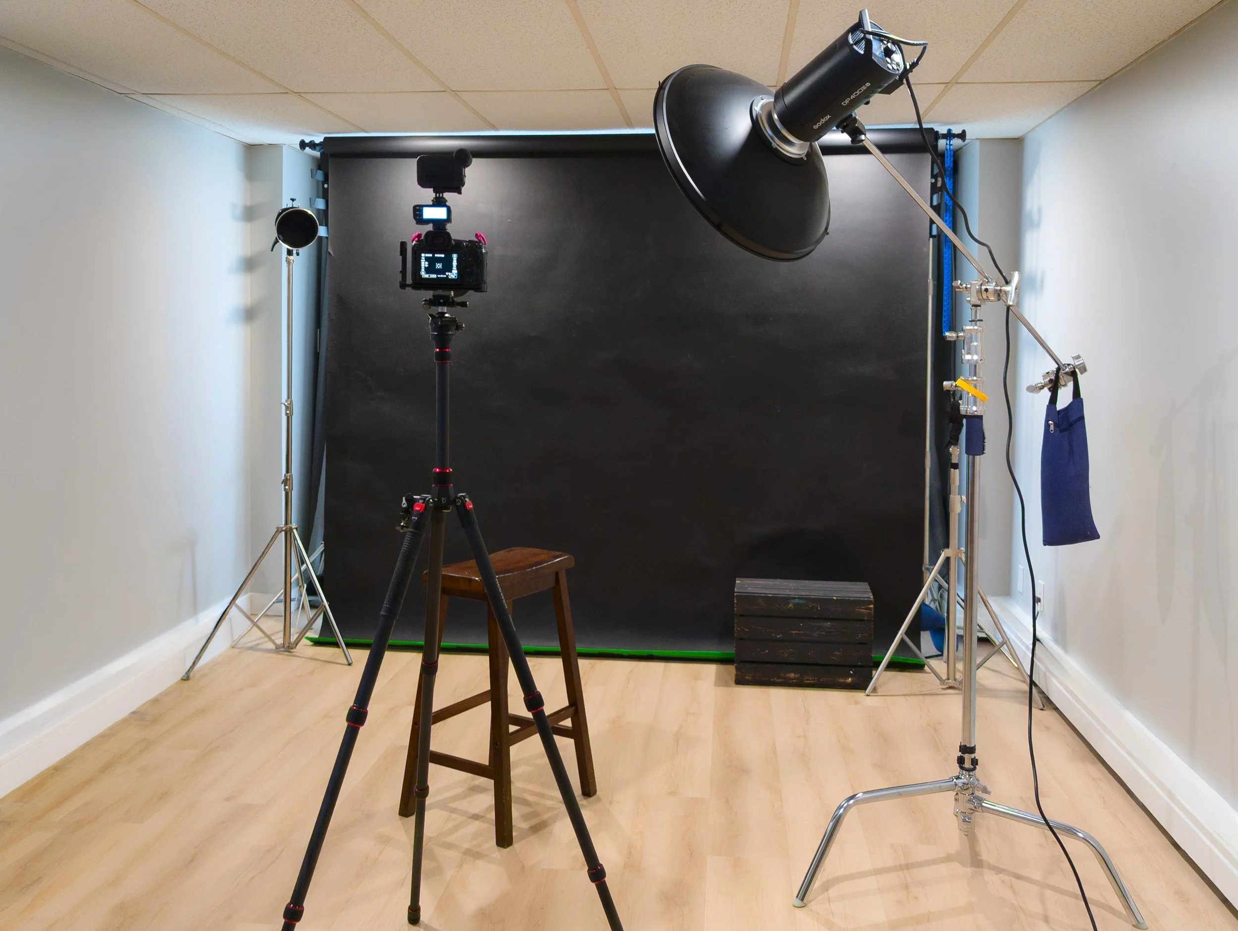 Photography studio setup with black backdrop, tripod-mounted camera, studio lighting equipment, a stool, and equipment stand in a room with hardwood floors and white walls.