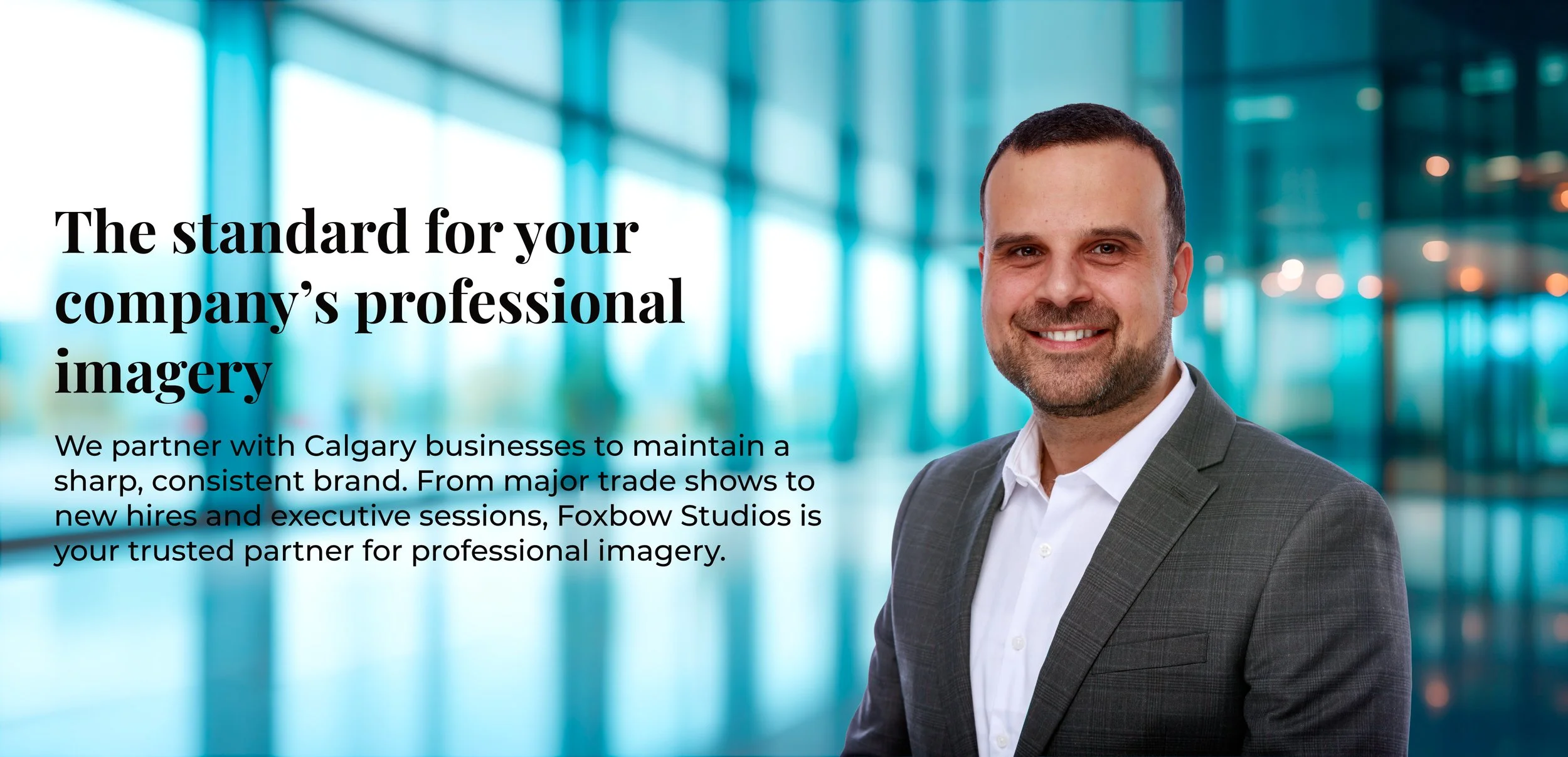 A professional man in a gray suit smiling in a modern office lobby with glass walls and bright lighting.