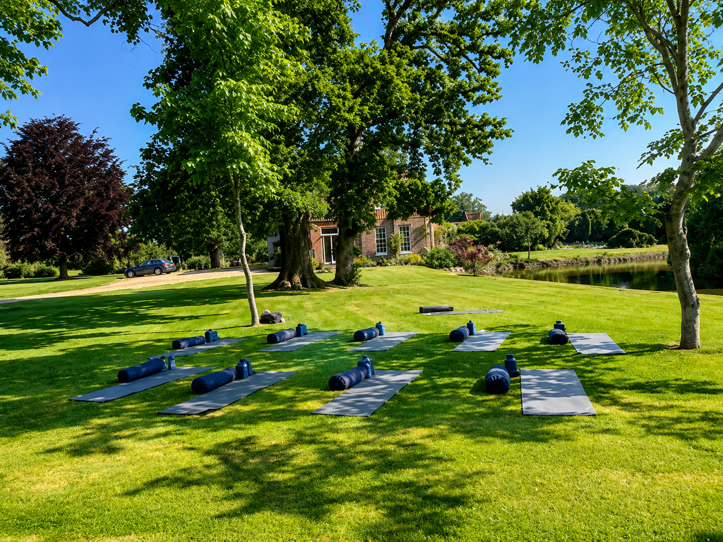 Outdoor yoga setup on grass with yoga mats and rollers under trees near a house, with lake and trees in the background on a sunny day.