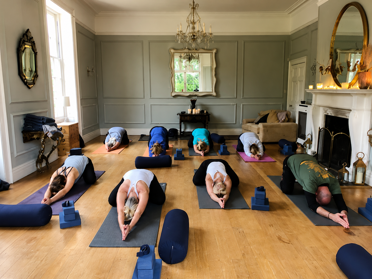 A group of people practicing yoga in a spacious, elegant living room with wooden floors, candles, and a fireplace, all positioned on yoga mats in a kneeling pose with hands clasped in prayer.