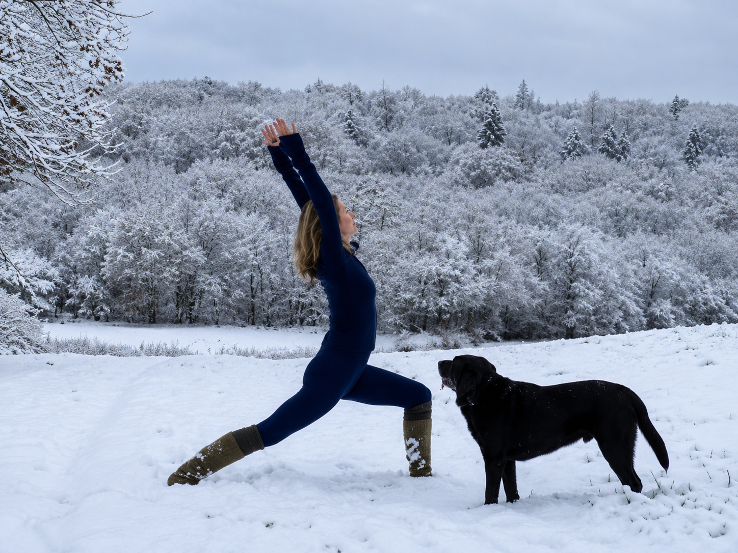 A woman practicing yoga outdoors in a snowy landscape with a black dog nearby, surrounded by snow-covered trees.