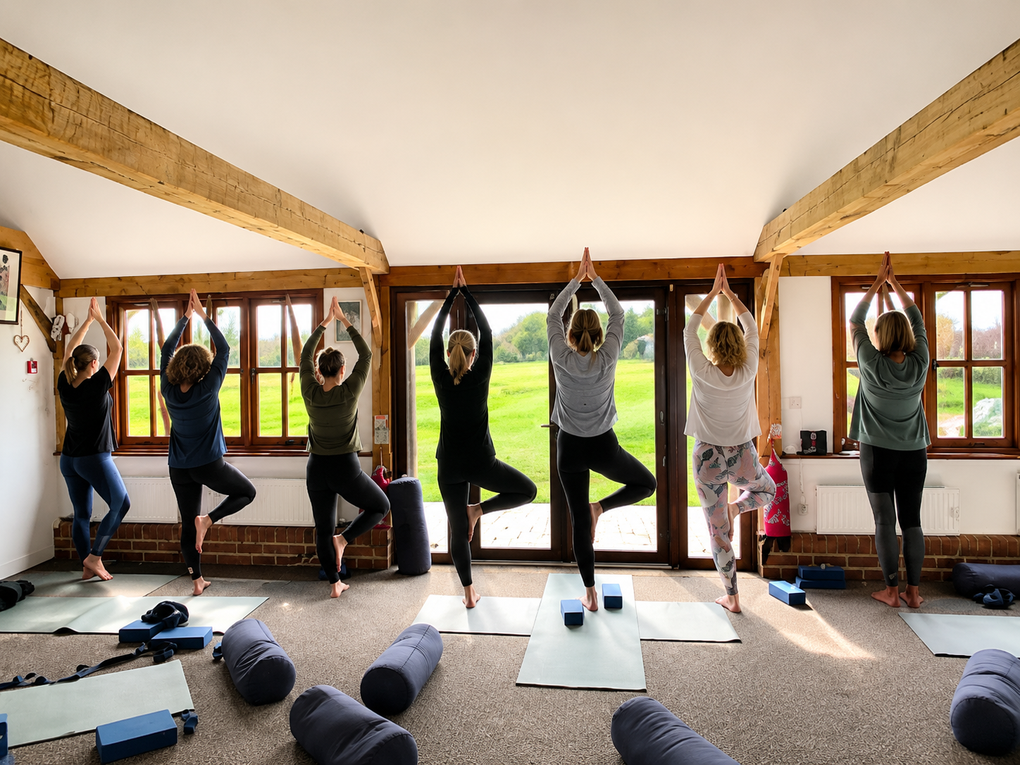 Group of women practicing yoga in a bright, rustic room with large windows overlooking a green field, all in tree pose with hands raised above heads.