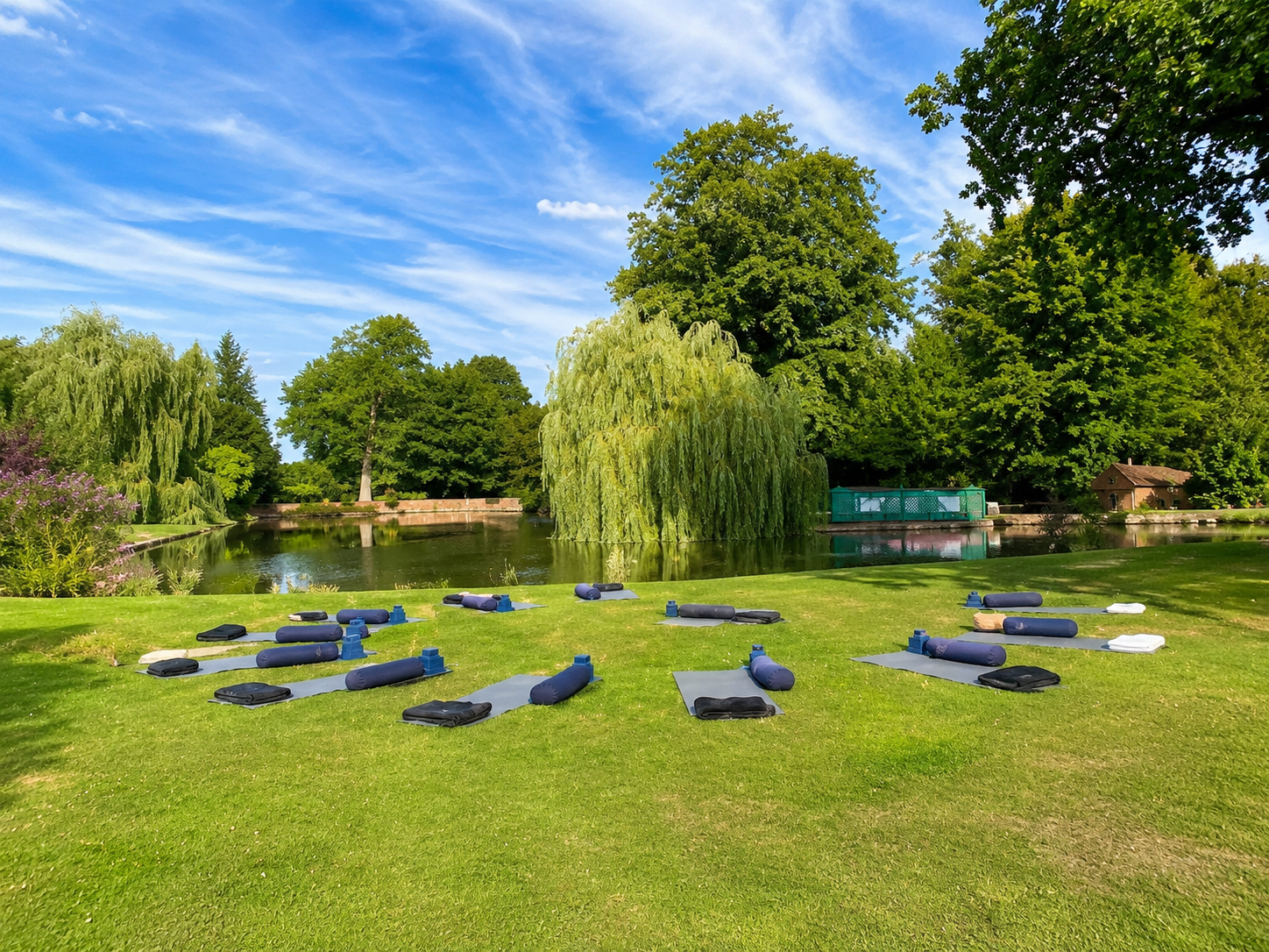 Yoga mats and props set up on a grassy area near a pond in a park, with trees and blue sky in the background.