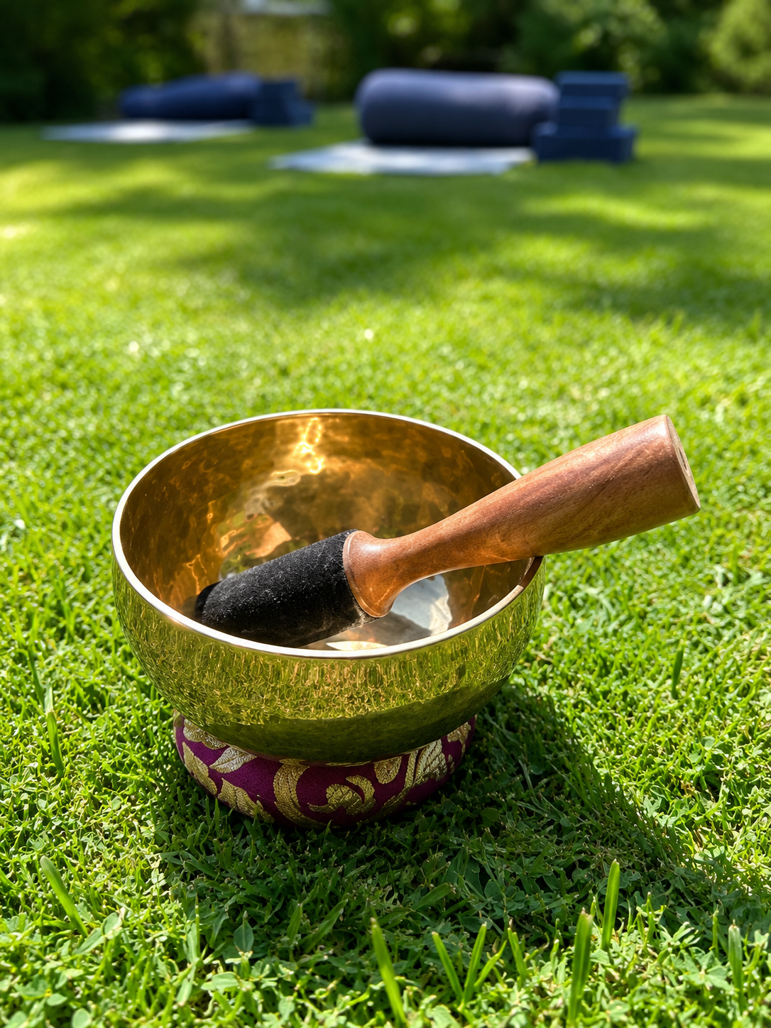 A brass singing bowl with a wooden striker resting inside, placed on a decorative purple cushion on green grass, with outdoor seating in the background.
