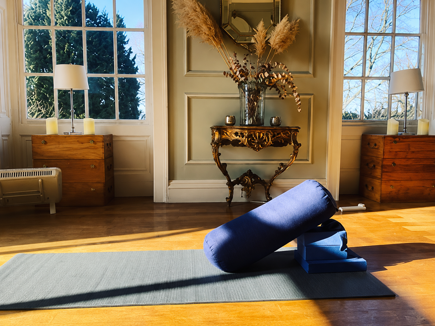 A yoga mat with a bolster and block in a daylight-lit, elegant room with large windows, wooden floors, and vintage furniture, featuring a decorative table with flower arrangements and candles.