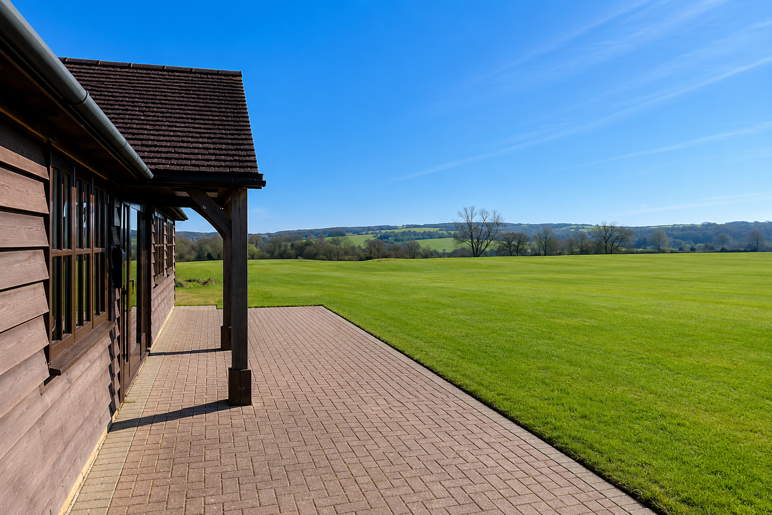 A wooden building with a sloped roof and large windows on a paved patio, overlooking a green field with trees and distant hills under a clear blue sky.