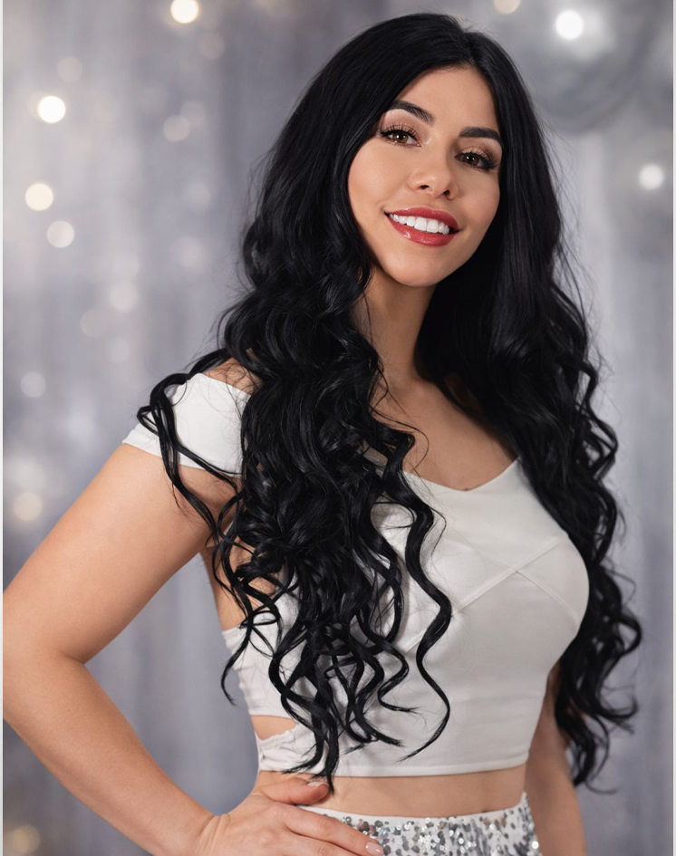 A woman with long, curly black hair, wearing a white crop top and a sparkly skirt, smiling with a gray, blurred background.