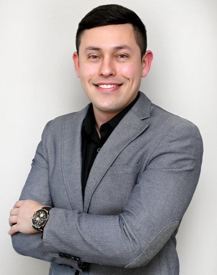 A young man with short dark hair, wearing a gray blazer over a black shirt, smiling with his arms crossed, standing against a plain white wall.