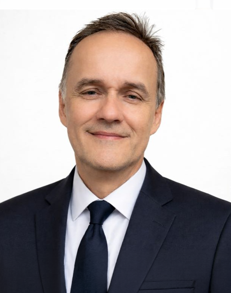 A middle-aged man with light skin, light brown hair, and blue eyes wearing a black suit, white shirt, and dark blue tie, smiling slightly against a plain white background.