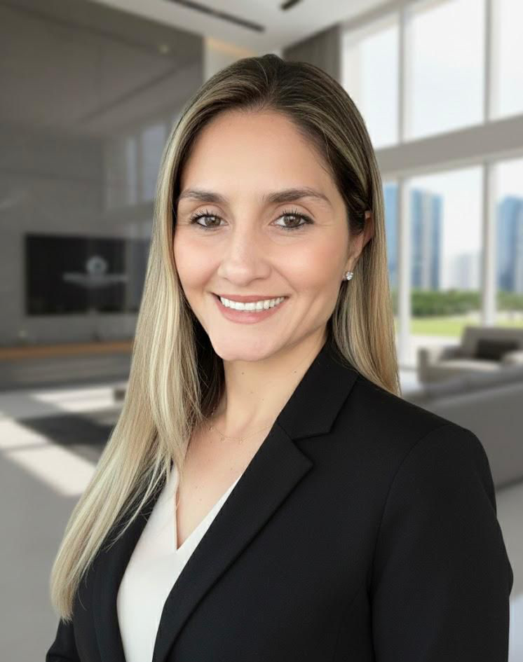 A woman with long blonde hair, wearing a black blazer and white top, smiling in a modern office with large windows and a cityscape view.