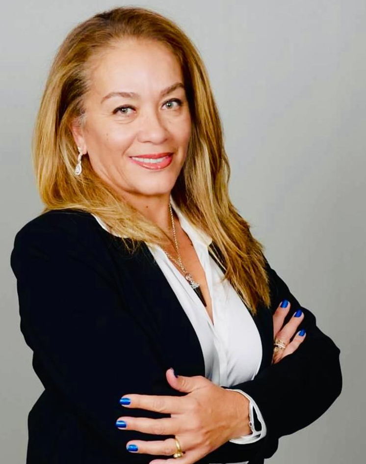 A woman with blond hair wearing a black blazer and white shirt, smiling with her arms crossed, against a plain gray background.