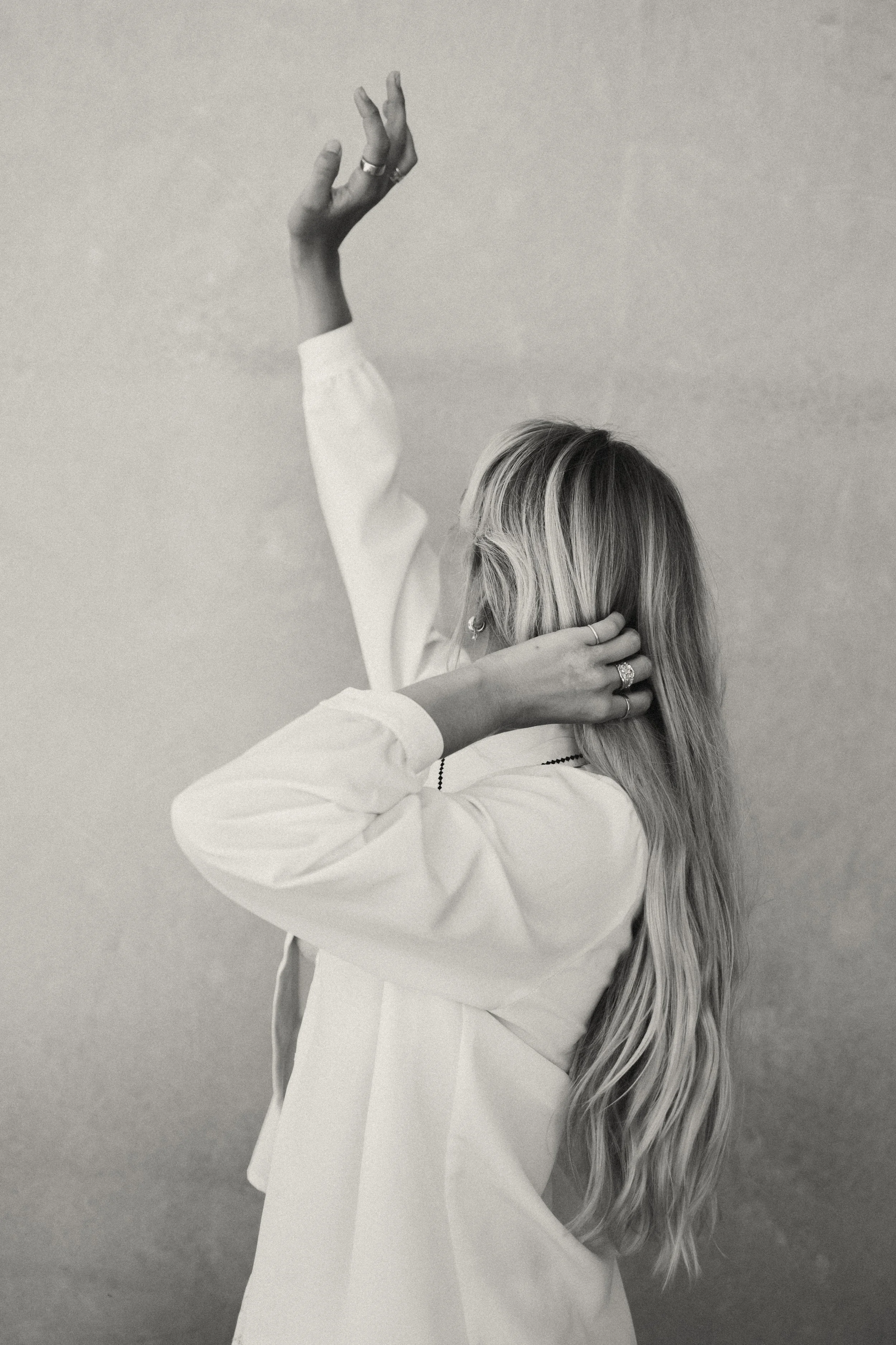 A woman with long, wavy hair adjusts her hair with one hand while her other arm is raised, against a plain background.