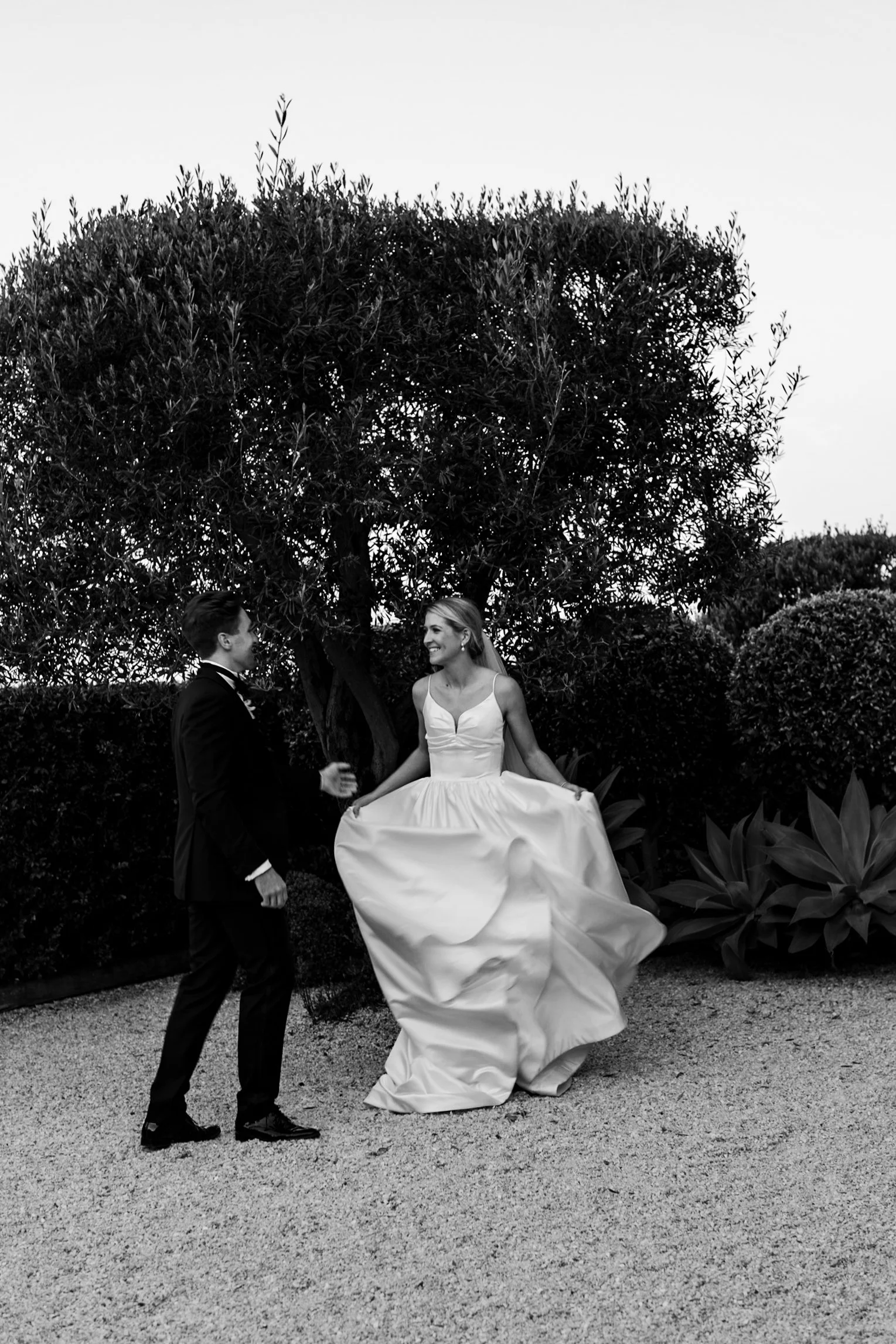 A black and white photo of a bride in a wedding dress smiling at a groom in a suit, standing outdoors on a gravel path near a large bush and trees.
