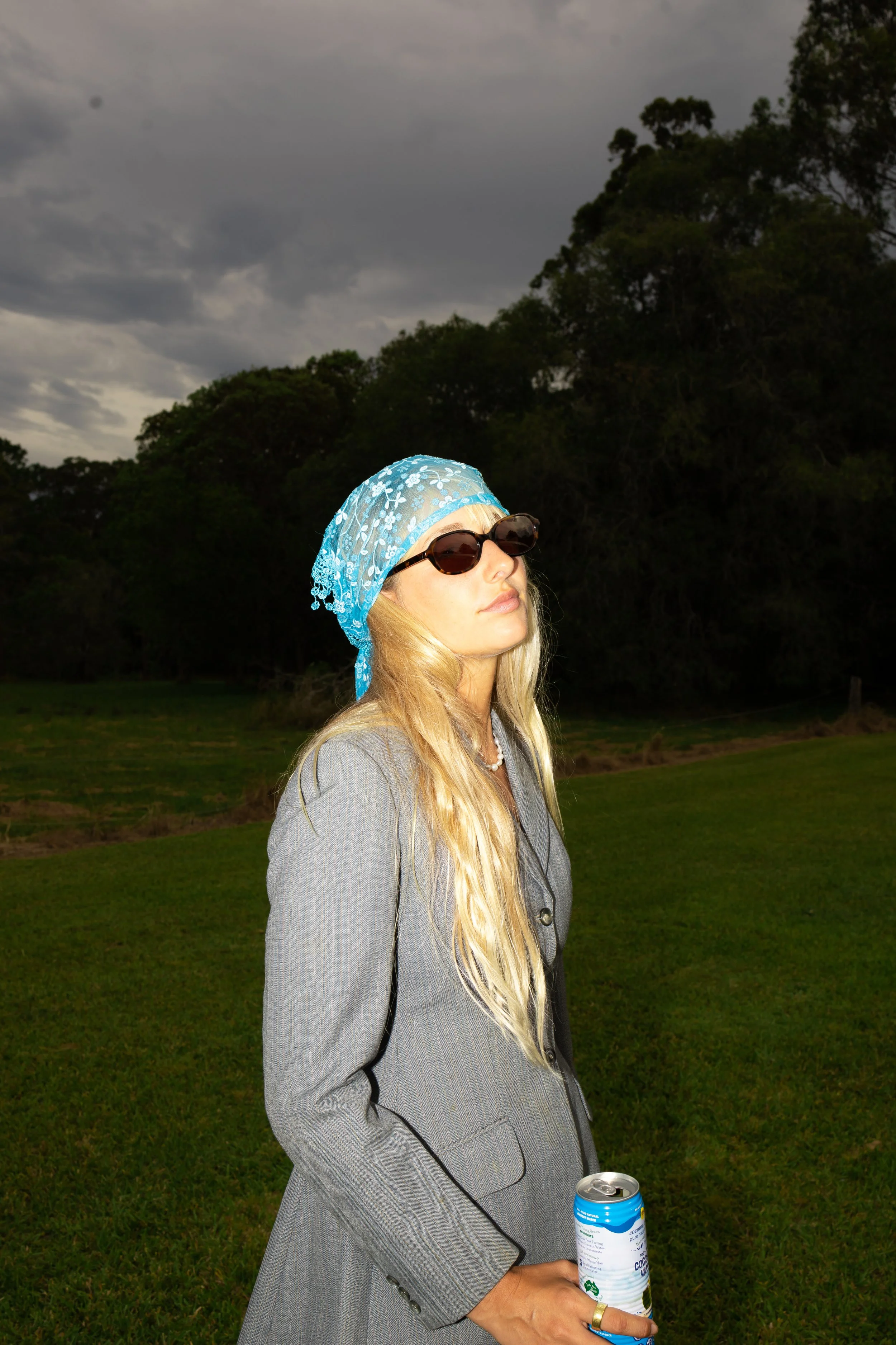 A woman with long blonde hair, wearing sunglasses, a gray blazer, a blue lace headscarf, and a pearl necklace, stands outdoors on a grassy area with dark clouds overhead. She is holding a can of soda in her right hand.