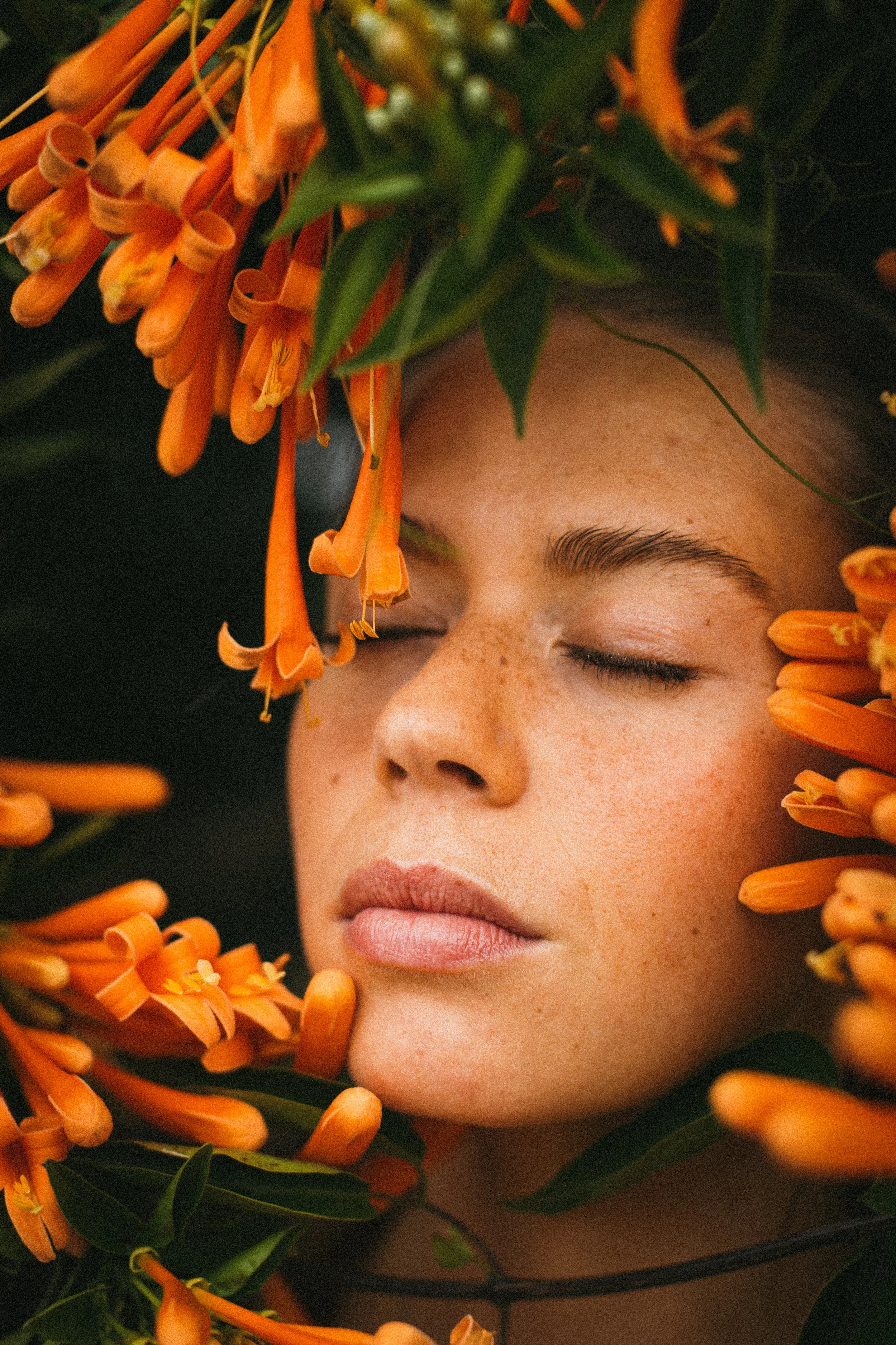 A young woman with closed eyes surrounded by orange flowers and green leaves.