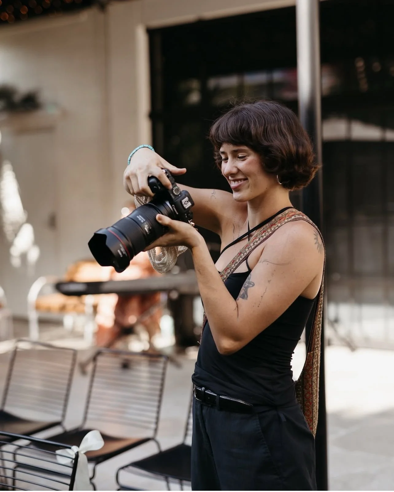 Photographer with short curly hair and tattoos on her arm looking at a camera and smiling outdoors.