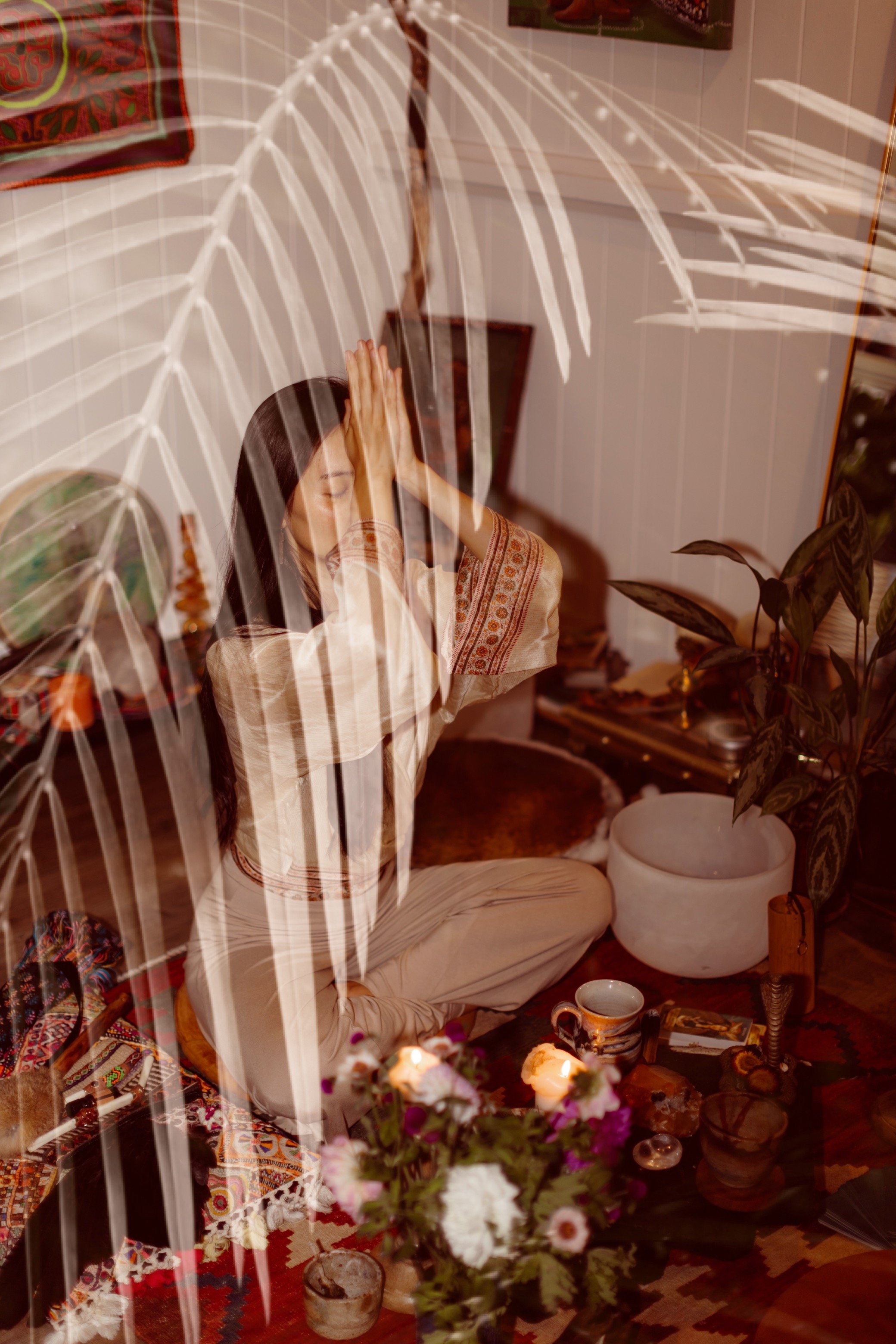 A woman practicing yoga or meditation indoors, sitting cross-legged with hands pressed together in prayer position, surrounded by candles, flowers, and spiritual objects, seen through a reflection of a large white-fingered leaf.