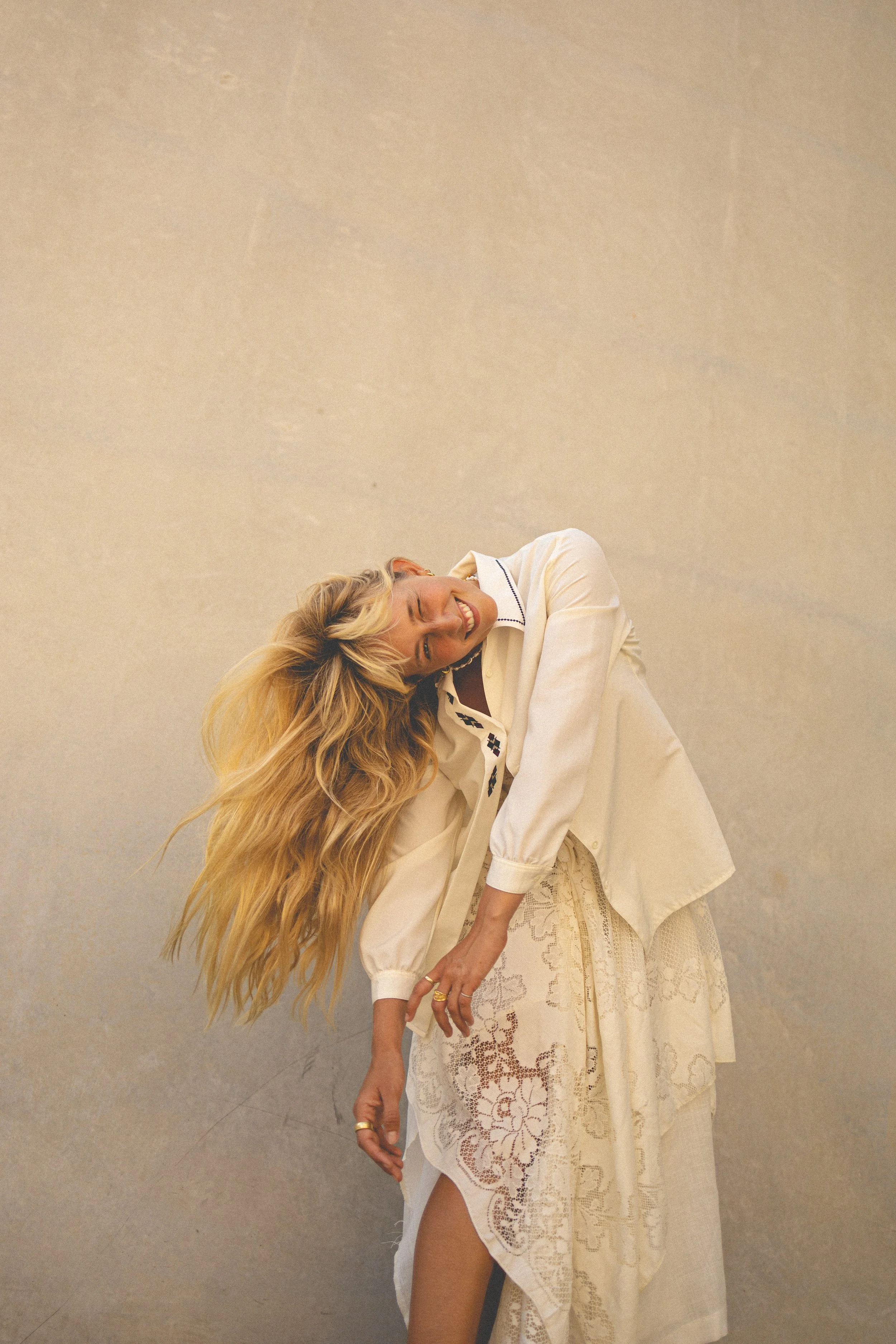 A woman with long blonde hair, smiling and leaning to the side, wearing a cream-colored blouse and a lace skirt, standing against a plain beige wall.