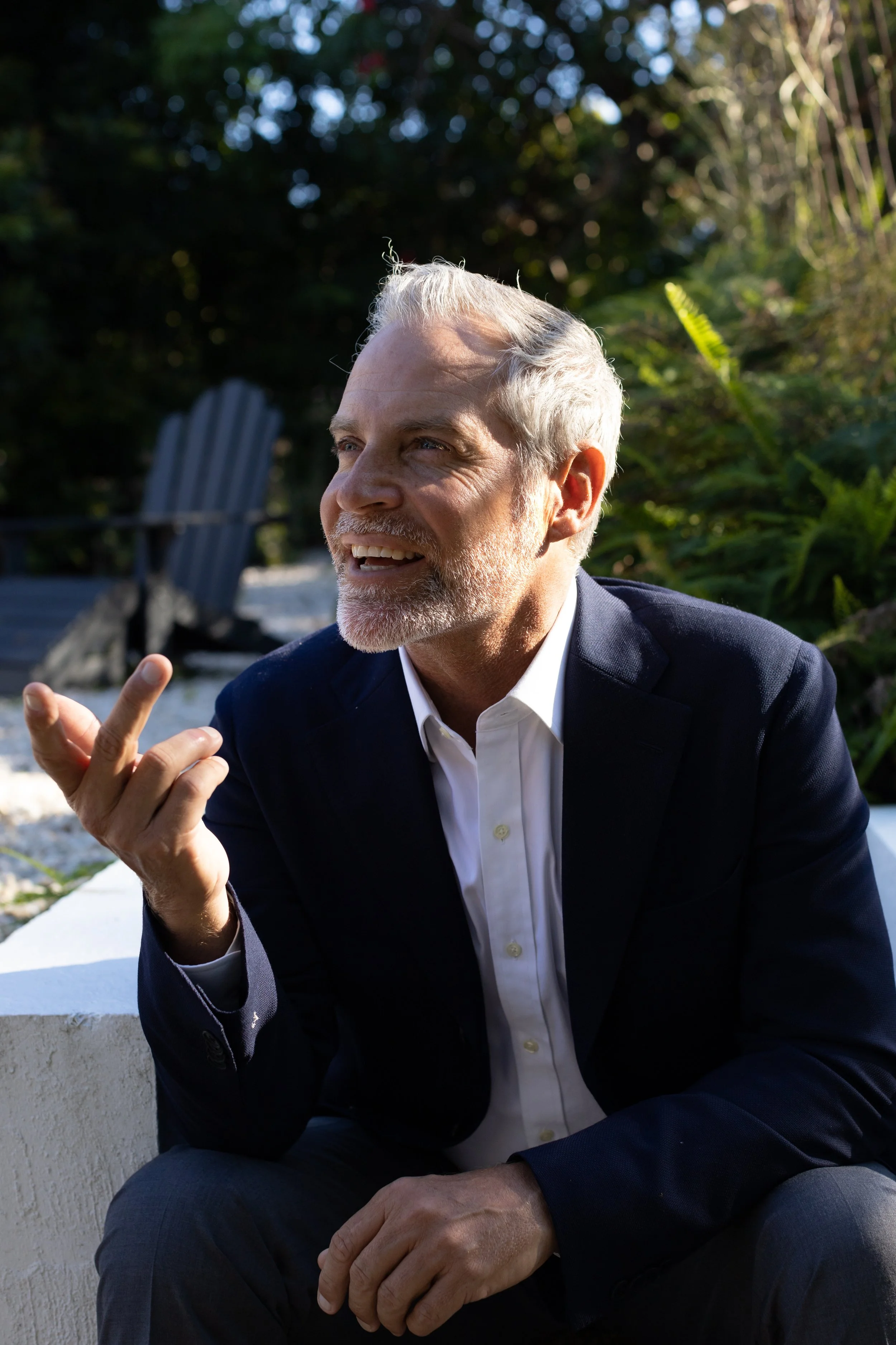 A mature man in a suit talking outdoors with greenery in the background.