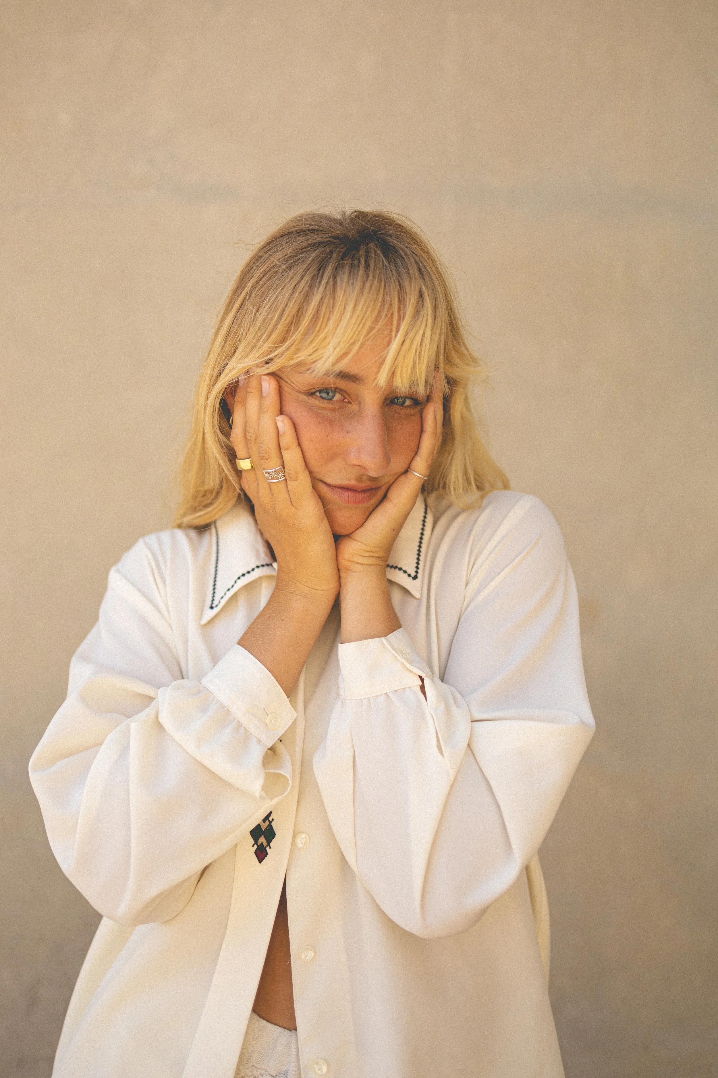 A young woman with blonde hair and blue eyes holding her face with both hands, wearing a white shirt with embroidered details, standing against a plain beige background.