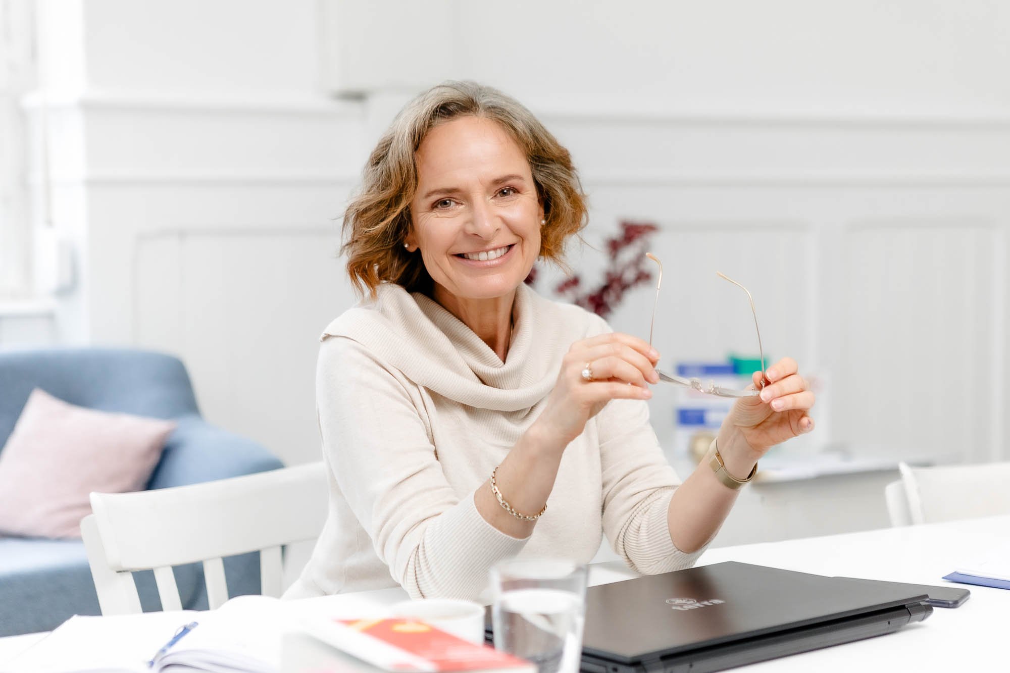 Frau mit Brille und Schmuck, lächelnd bei einem Treffen im Büro, sitzend an einem weißen Tisch mit Laptop, Kaffee, Dokumenten und einem Notizbuch.