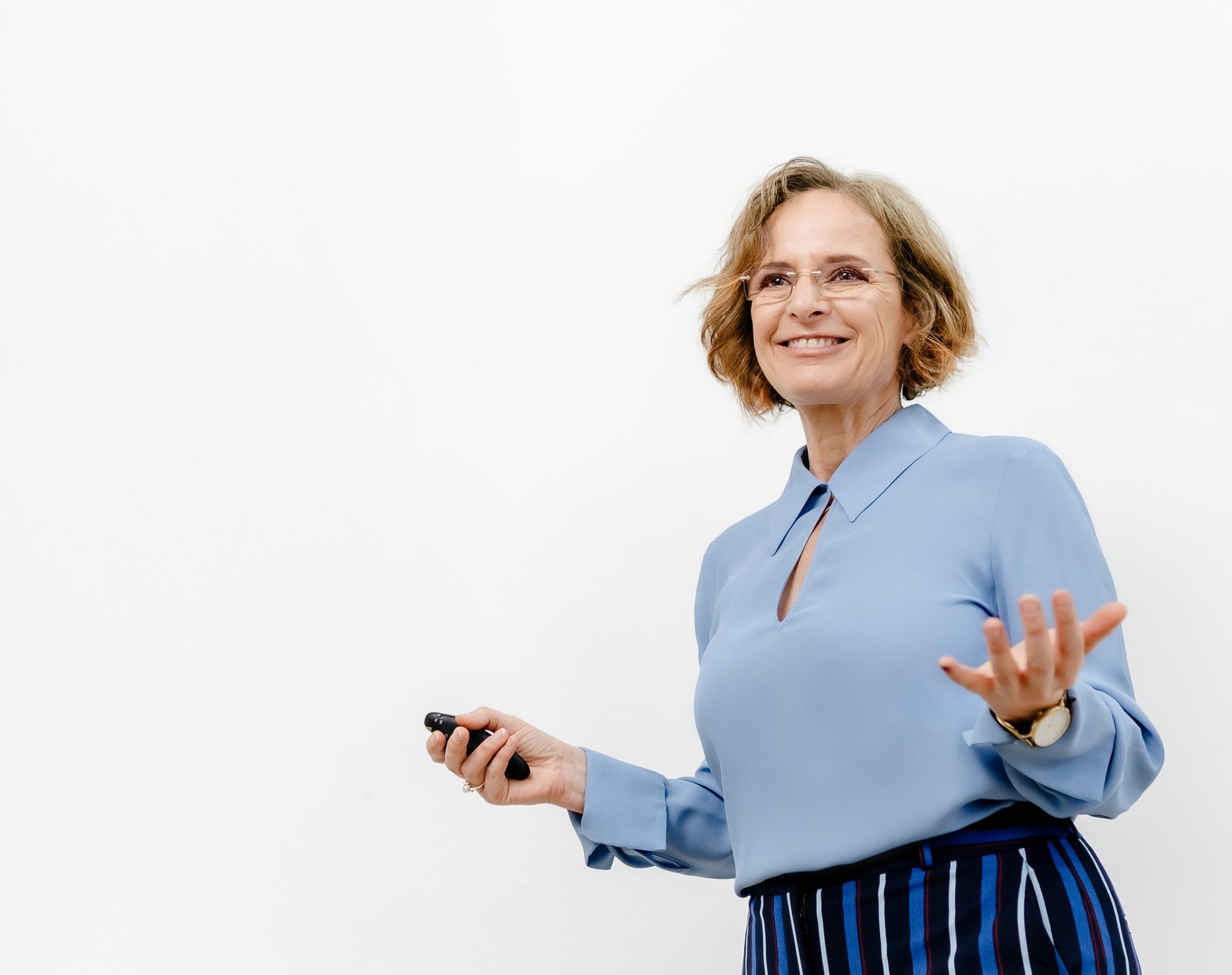 Eine lachende Frau in blauer Bluse hält eine Fernbedienung in der rechten Hand und gestikuliert mit der linken Hand.