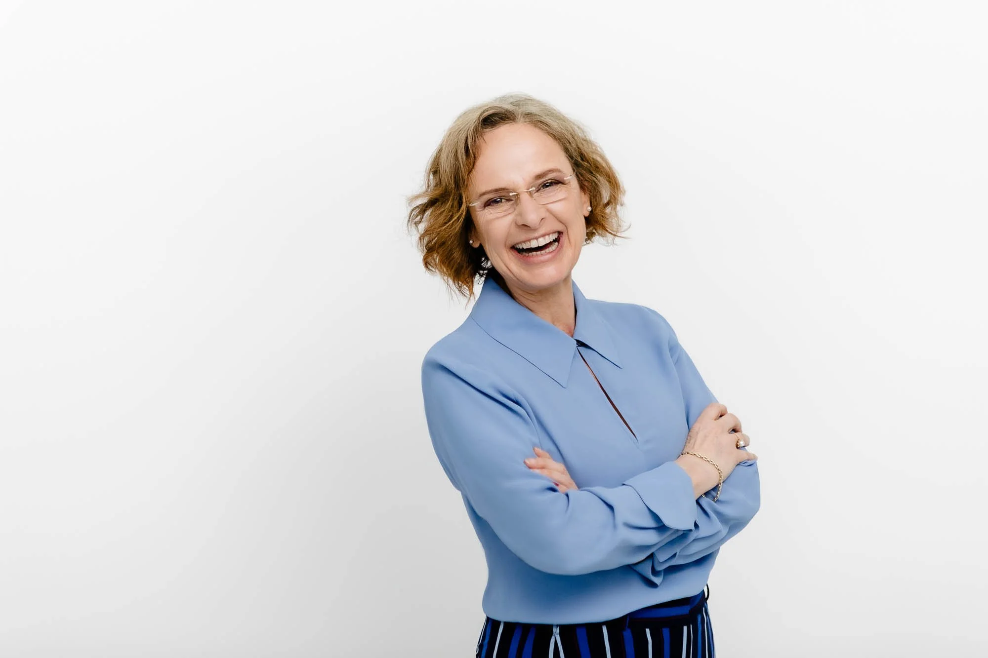 Lächelnde Frau mit rotbraunen, lockigen Haaren, Brille und blauer Bluse, vor weißem Hintergrund.