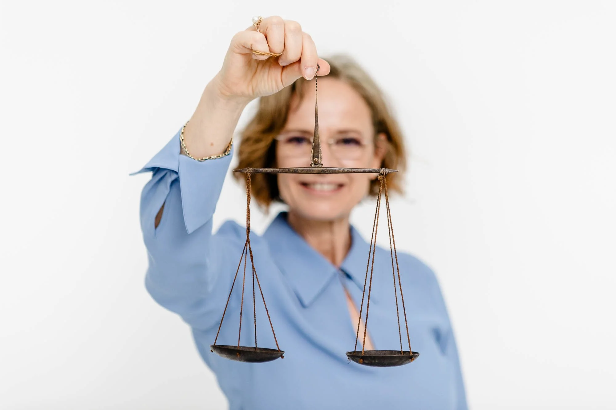 Eine Frau hält eine alte, rostige Waage in der Hand vor einem weißen Hintergrund, lächelt und trägt eine blaue Bluse.